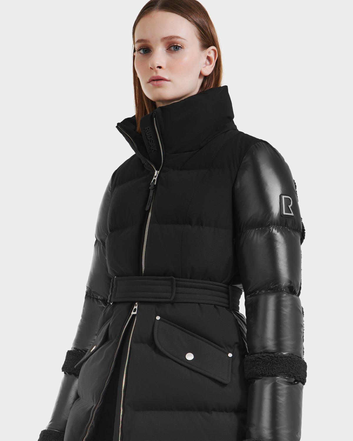 A woman models the Rudsak AUDREY L - 8124585 Black/Black, a knee-length puffer jacket with RDS certified down fill and shiny sleeves, standing confidently against a plain white background.