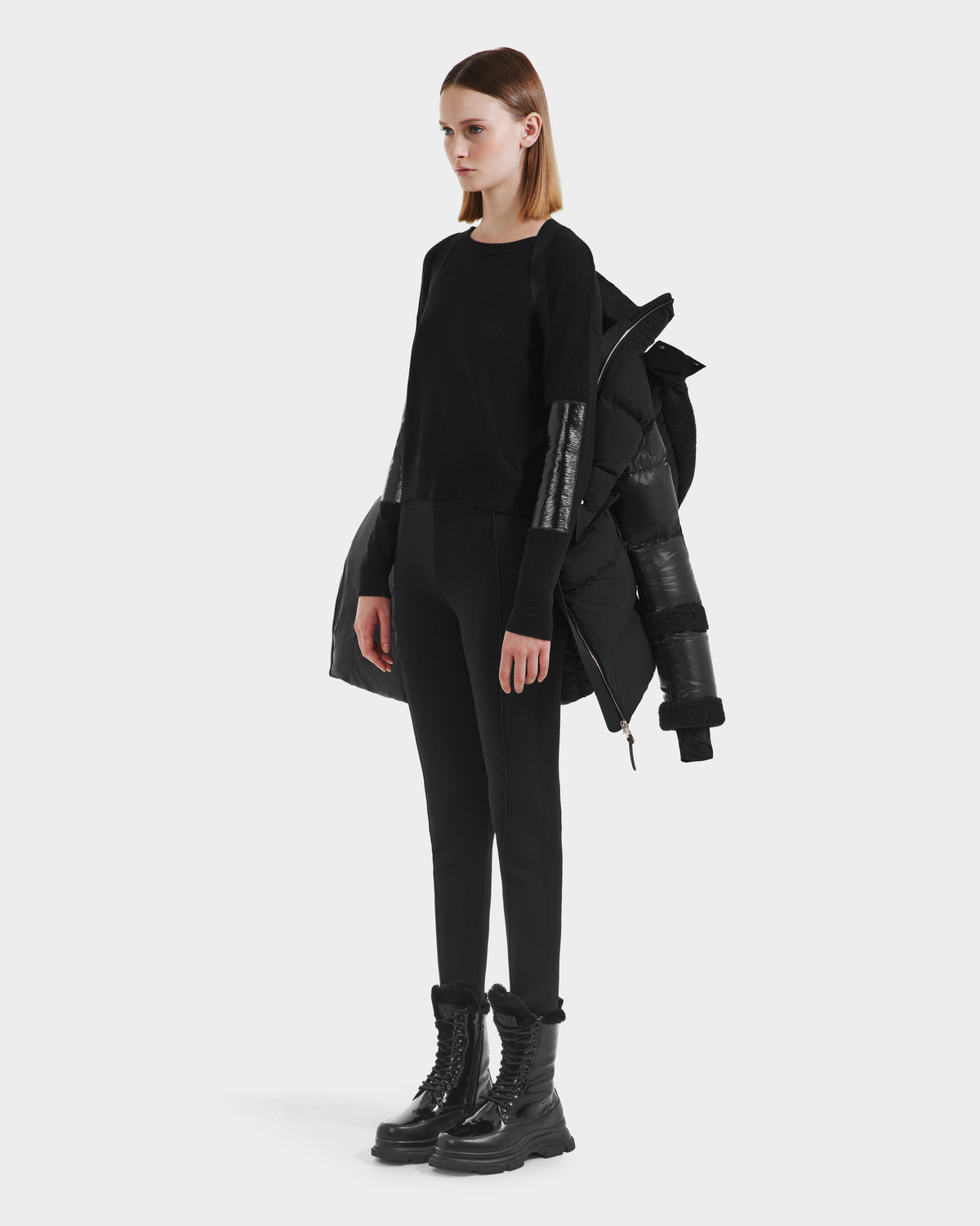 A woman faces left against a plain white background, wearing the Rudsak AUDREY L - 8124585 Black/Black knee-length down puffer jacket draped over her shoulders, styled with a black long-sleeve top, black pants, and chunky boots.