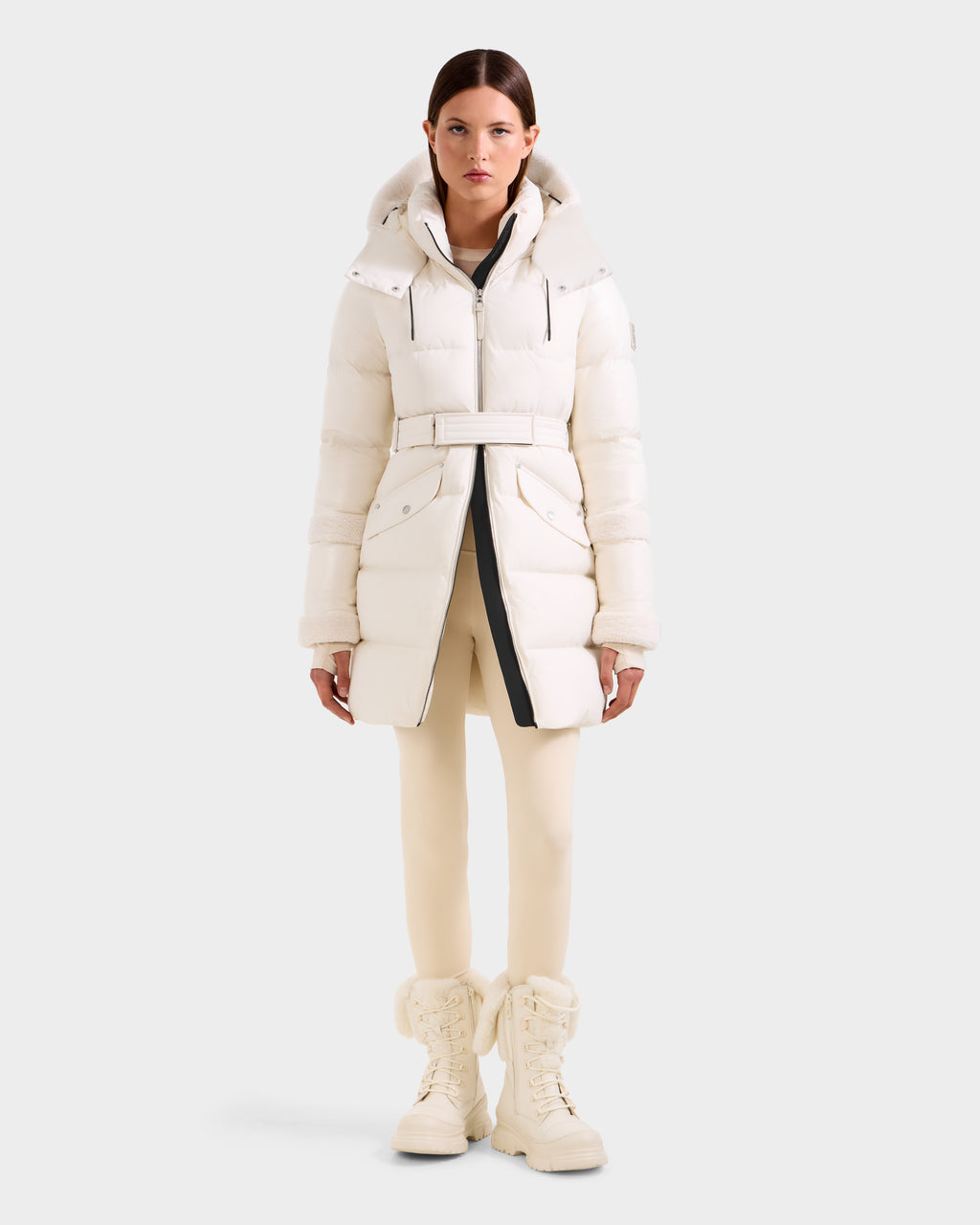 A woman faces forward in the Rudsak AUDREY L - 8124585 Vanilla knee-length, off-white down puffer made from recycled Stormshell fabric and RDS certified down, featuring a belt, hood, pockets; styled with matching leggings and tall lace-up boots.