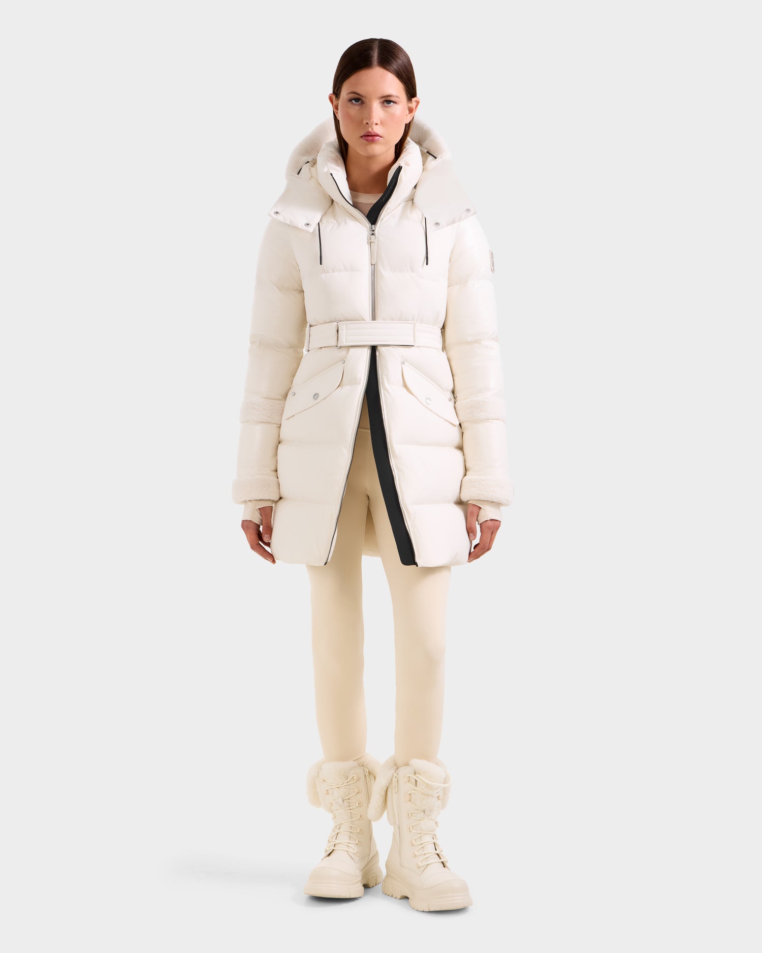 A woman faces forward in the Rudsak AUDREY L - 8124585 Vanilla knee-length, off-white down puffer made from recycled Stormshell fabric and RDS certified down, featuring a belt, hood, pockets; styled with matching leggings and tall lace-up boots.