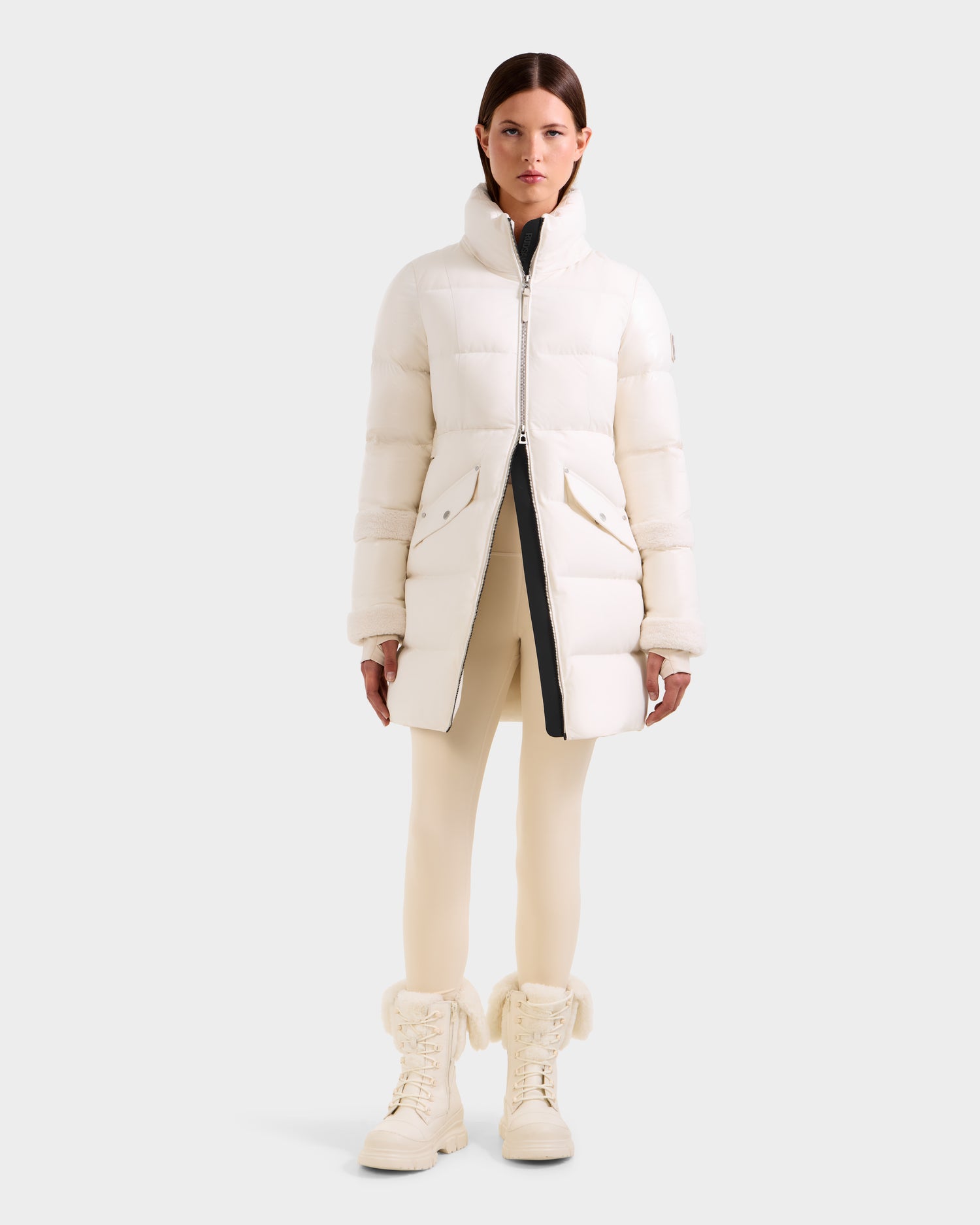A woman faces forward in a knee-length Rudsak AUDREY L - 8124585 Vanilla down puffer jacket, crafted from recycled Stormshell fabric with RDS certified down, paired with cream leggings and white fur-trimmed boots against a light gray backdrop.