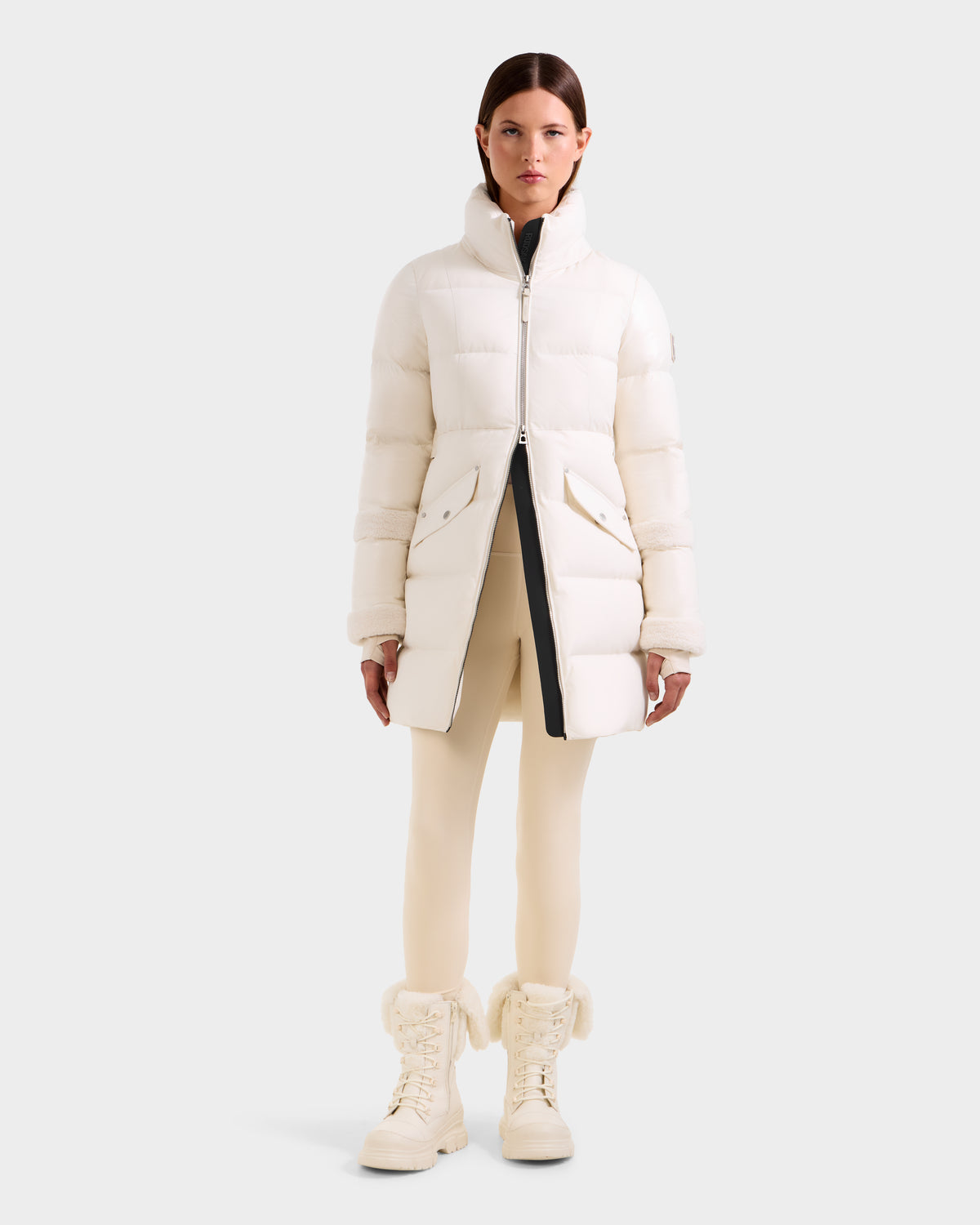 A woman faces forward in a knee-length Rudsak AUDREY L - 8124585 Vanilla down puffer jacket, crafted from recycled Stormshell fabric with RDS certified down, paired with cream leggings and white fur-trimmed boots against a light gray backdrop.