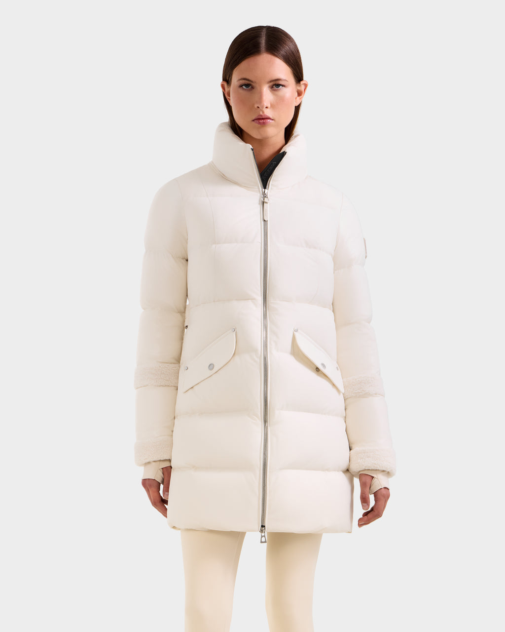 A woman models the Rudsak AUDREY L - 8124585 Vanilla knee-length down puffer jacket with a high collar and large front pockets, standing against a plain light gray background.