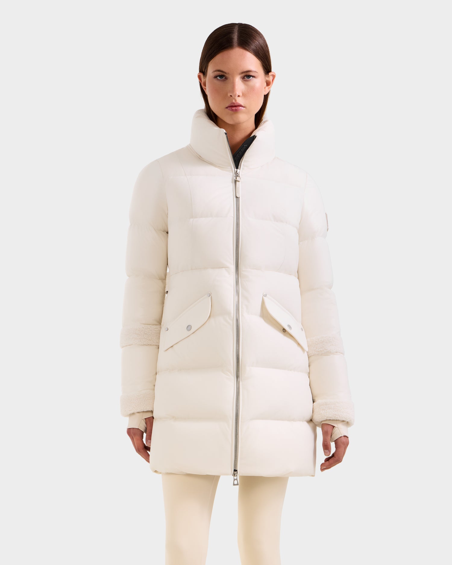 A woman models the Rudsak AUDREY L - 8124585 Vanilla knee-length down puffer jacket with a high collar and large front pockets, standing against a plain light gray background.