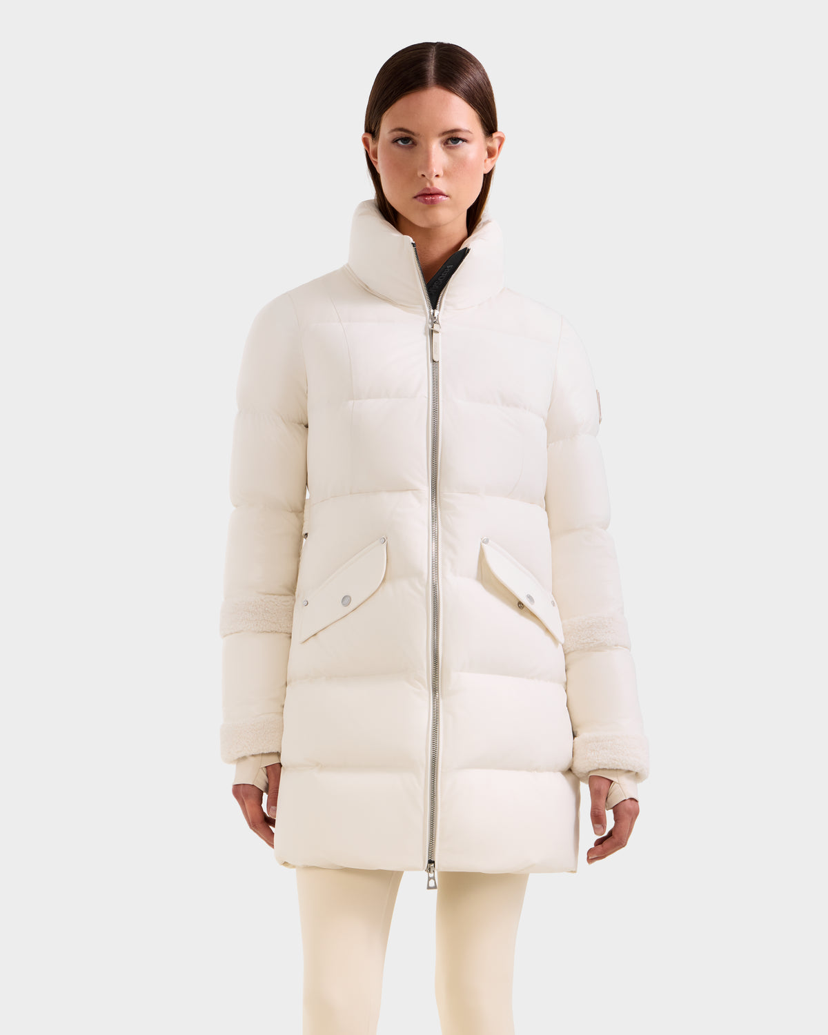 A woman models the Rudsak AUDREY L - 8124585 Vanilla knee-length down puffer jacket with a high collar and large front pockets, standing against a plain light gray background.