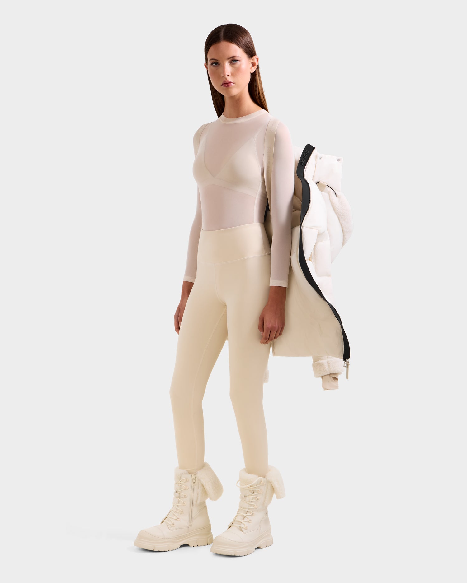 A woman poses confidently in Rudsak's AUDREY L - 8124585 Vanilla, draping the knee-length down puffer jacket over one shoulder. She pairs it with ivory leggings, a sheer long-sleeve top, and white boots against a plain background.
