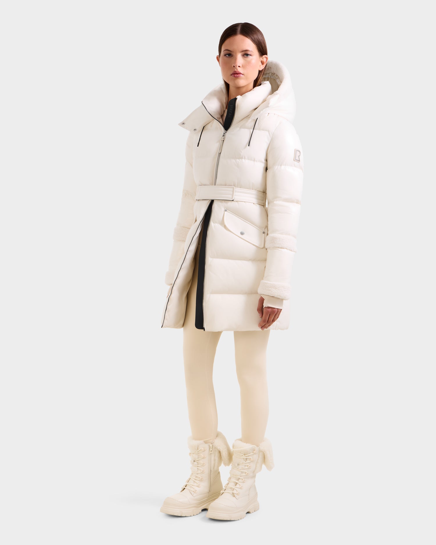 A woman faces forward in a Rudsak AUDREY L - 8124585 Vanilla outfit: an all-white, knee-length down puffer jacket with recycled Stormshell fabric and RDS certified down, matching skinny pants, and lace-up boots against a light gray background.