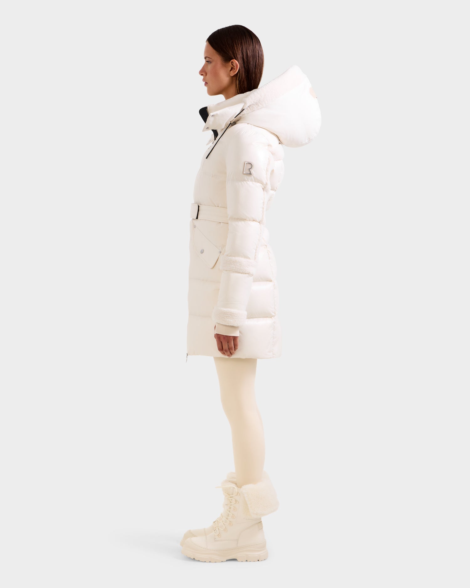 A woman in profile wears the Rudsak AUDREY L - 8124585 Vanilla knee-length puffer jacket with a hood, made from recycled Stormshell and filled with RDS certified down, paired with light leggings and fur-trimmed winter boots against a plain background.