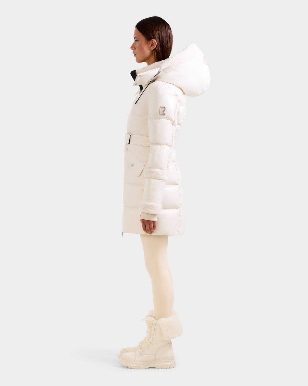 A woman in profile wears the Rudsak AUDREY L - 8124585 Vanilla knee-length puffer jacket with a hood, made from recycled Stormshell and filled with RDS certified down, paired with light leggings and fur-trimmed winter boots against a plain background.