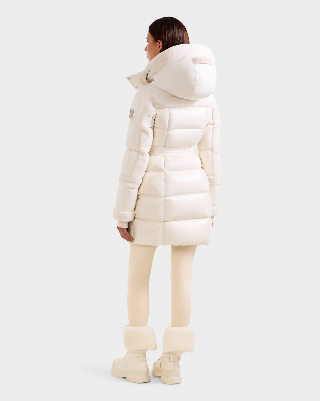 A woman stands facing away in the Rudsak AUDREY L - 8124585 Vanilla knee-length down puffer jacket, crafted from recycled Stormshell fabric with RDS certified down, paired with cream leggings and white fur-trimmed boots against a light gray backdrop.