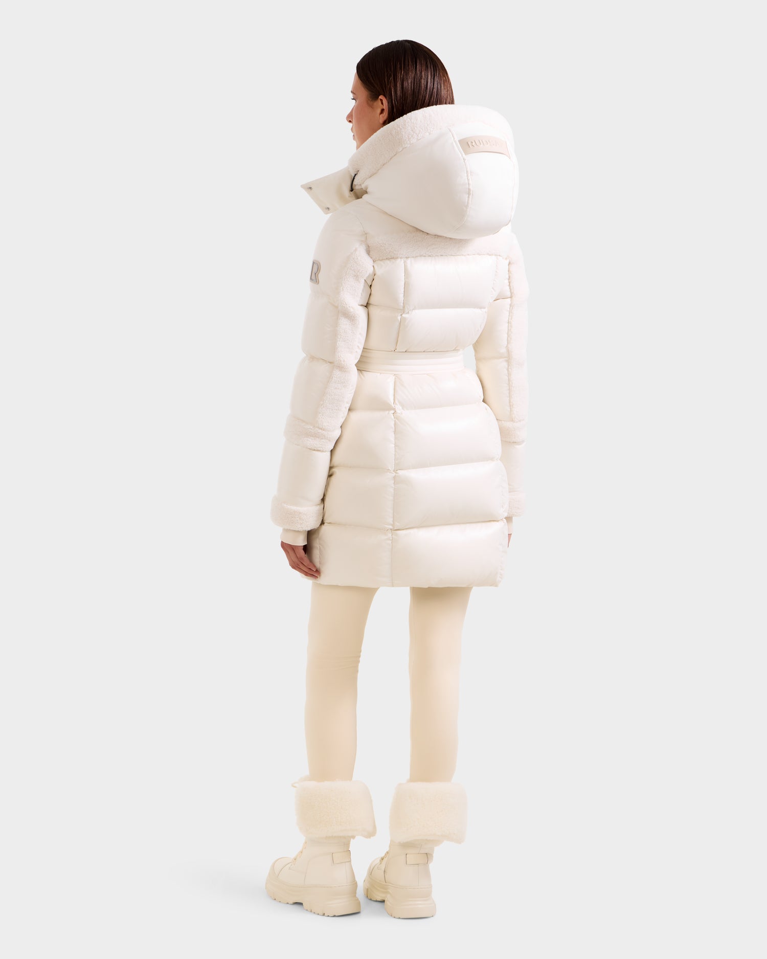 A woman stands facing away in the Rudsak AUDREY L - 8124585 Vanilla knee-length down puffer jacket, crafted from recycled Stormshell fabric with RDS certified down, paired with cream leggings and white fur-trimmed boots against a light gray backdrop.