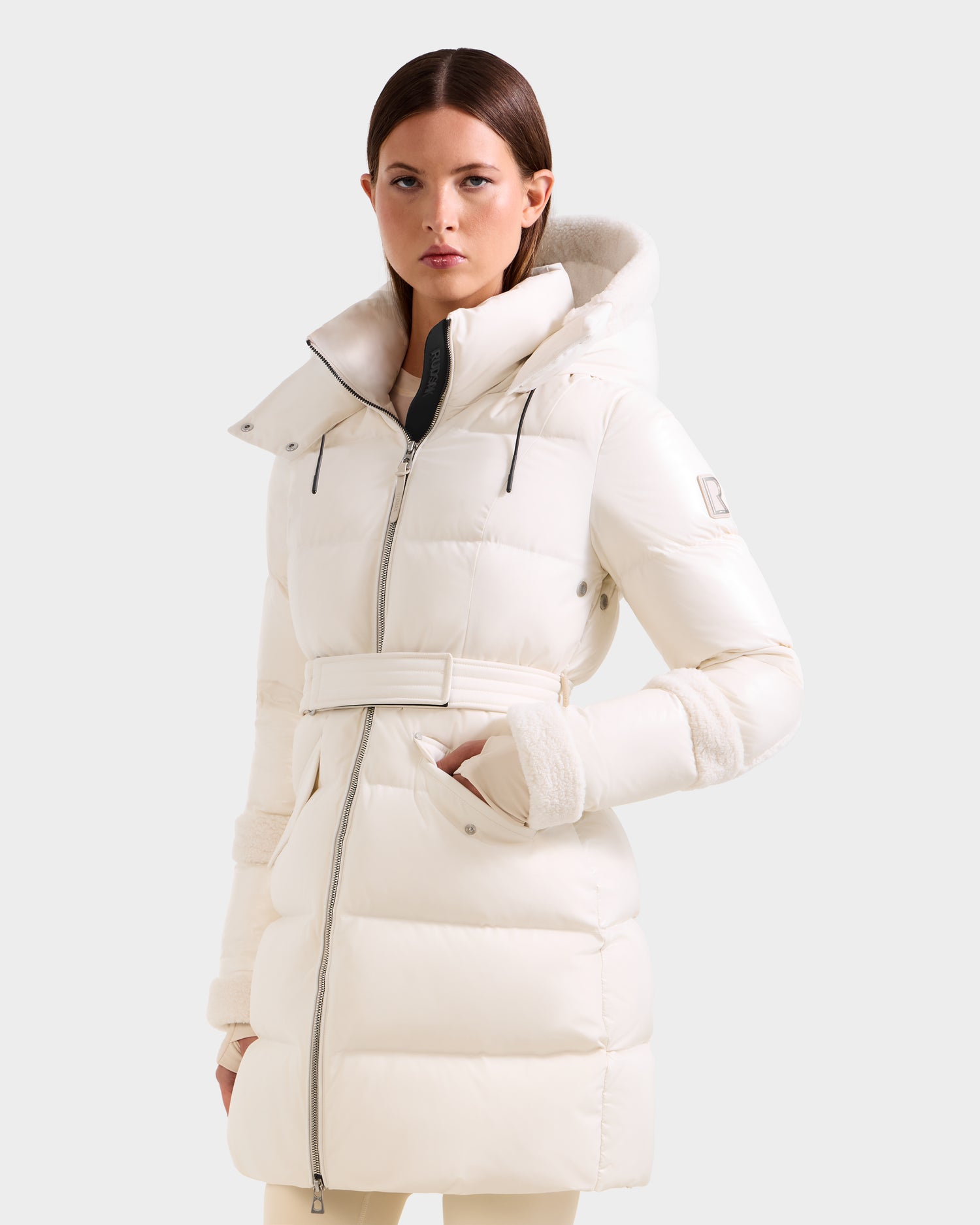 A woman with straight brown hair and a neutral expression models the Rudsak AUDREY L - 8124585 Vanilla, a knee-length down puffer jacket with high collar, belt, hood, and RDS certified filling, standing against a light gray background.
