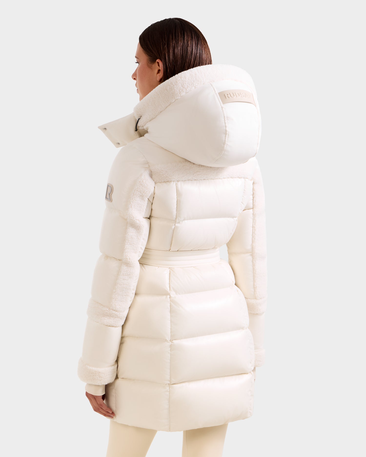 A woman with straight brown hair models the Rudsak AUDREY L - 8124585 Vanilla knee-length down puffer jacket with a large hood and plush detailing, made from recycled Stormshell fabric, shown from the back against a light gray background.