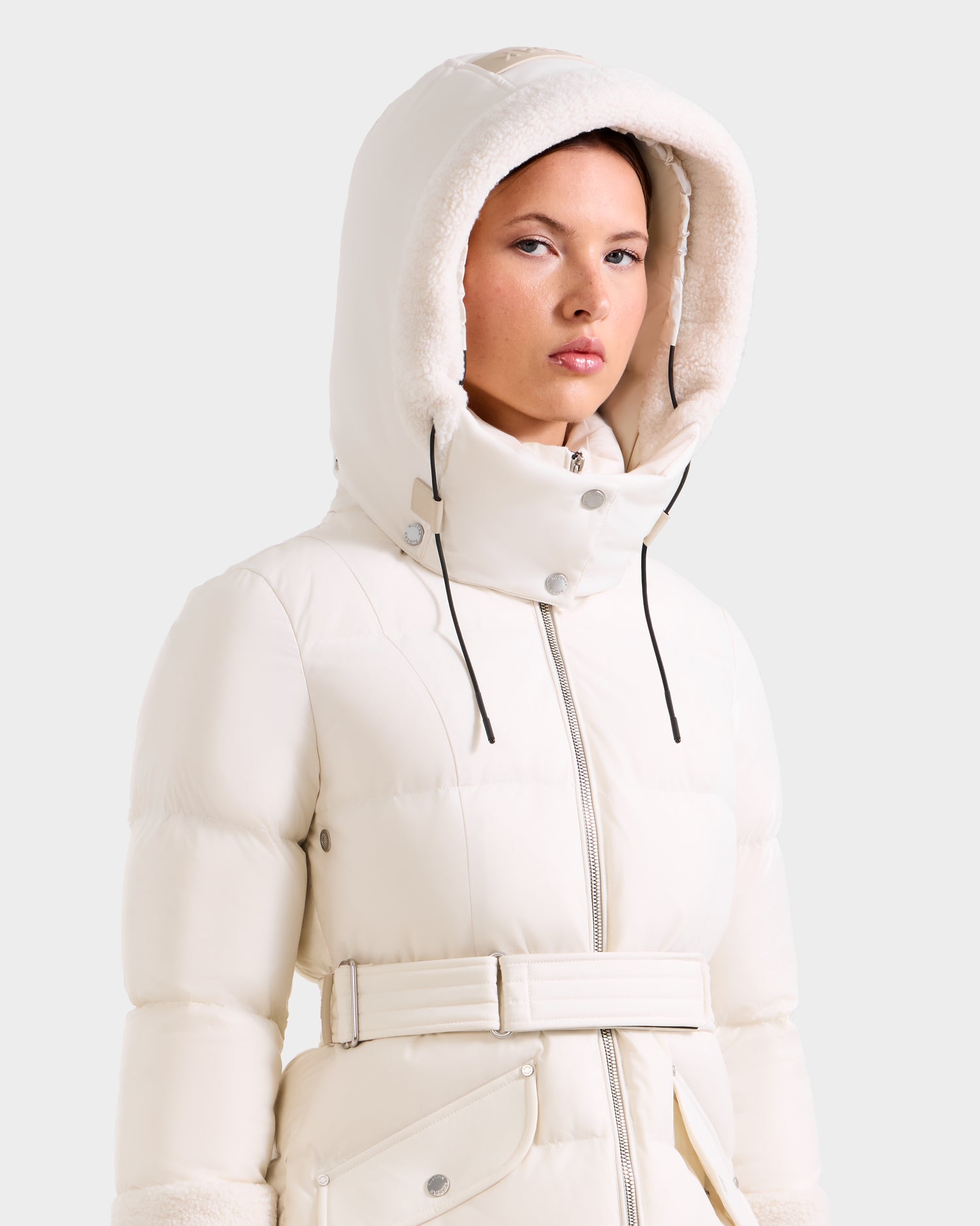 A woman wears the Rudsak AUDREY L - 8124585 Vanilla, a knee-length down puffer with a belted waist, faux fur hood trim, recycled Stormshell fabric, RDS certified down fill, front zipper, snap buttons, and adjustable hood drawstrings.