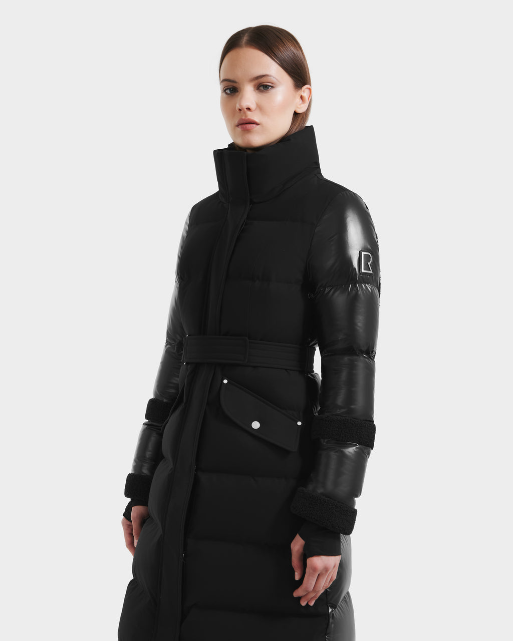 A woman models the Rudsak AUDREY XL - 8125601 Black/Black maxi-length down puffer jacket with a high collar and patch details, standing confidently against a plain white background.