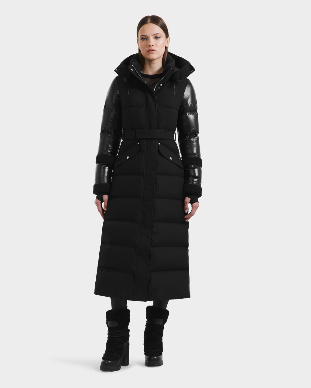 A woman faces forward in the Rudsak AUDREY XL - 8125601 Black/Black maxi down puffer, featuring a belted waist and shiny black sleeves. The coat, made from recycled Stormshell fabric, is paired with black boots against a plain white background.