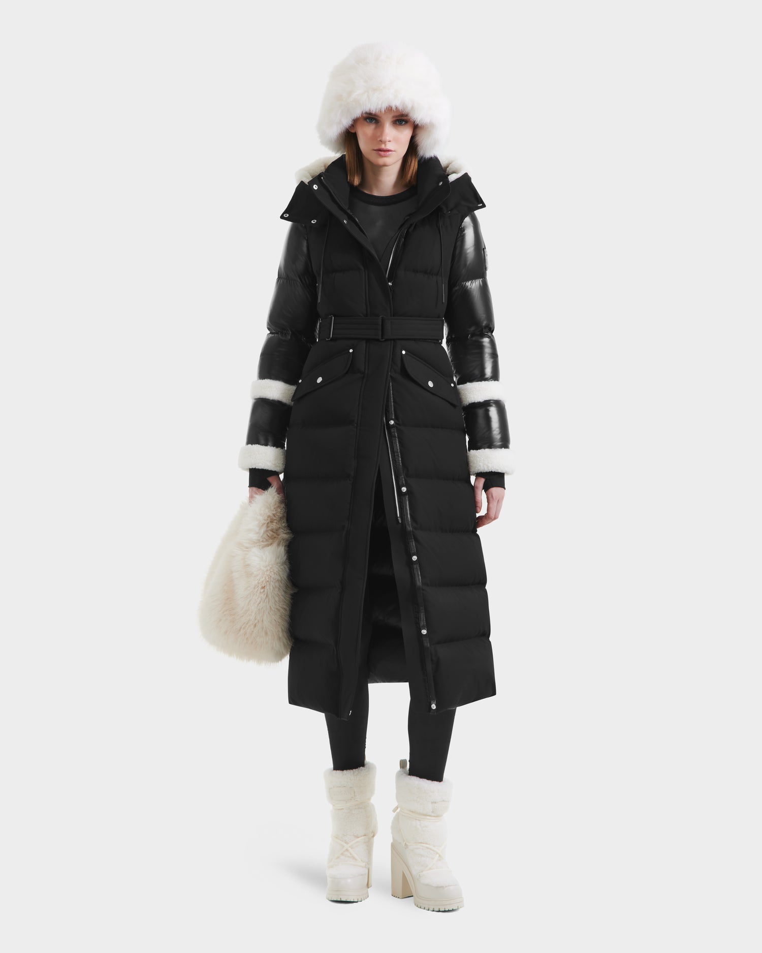 A woman wears the Rudsak AUDREY XL - 8125601 Black/Iceland maxi puffer with black leather sleeves and white faux shearling cuffs, paired with a white fur hat, boots, and a fluffy white bag, standing against a plain background.
