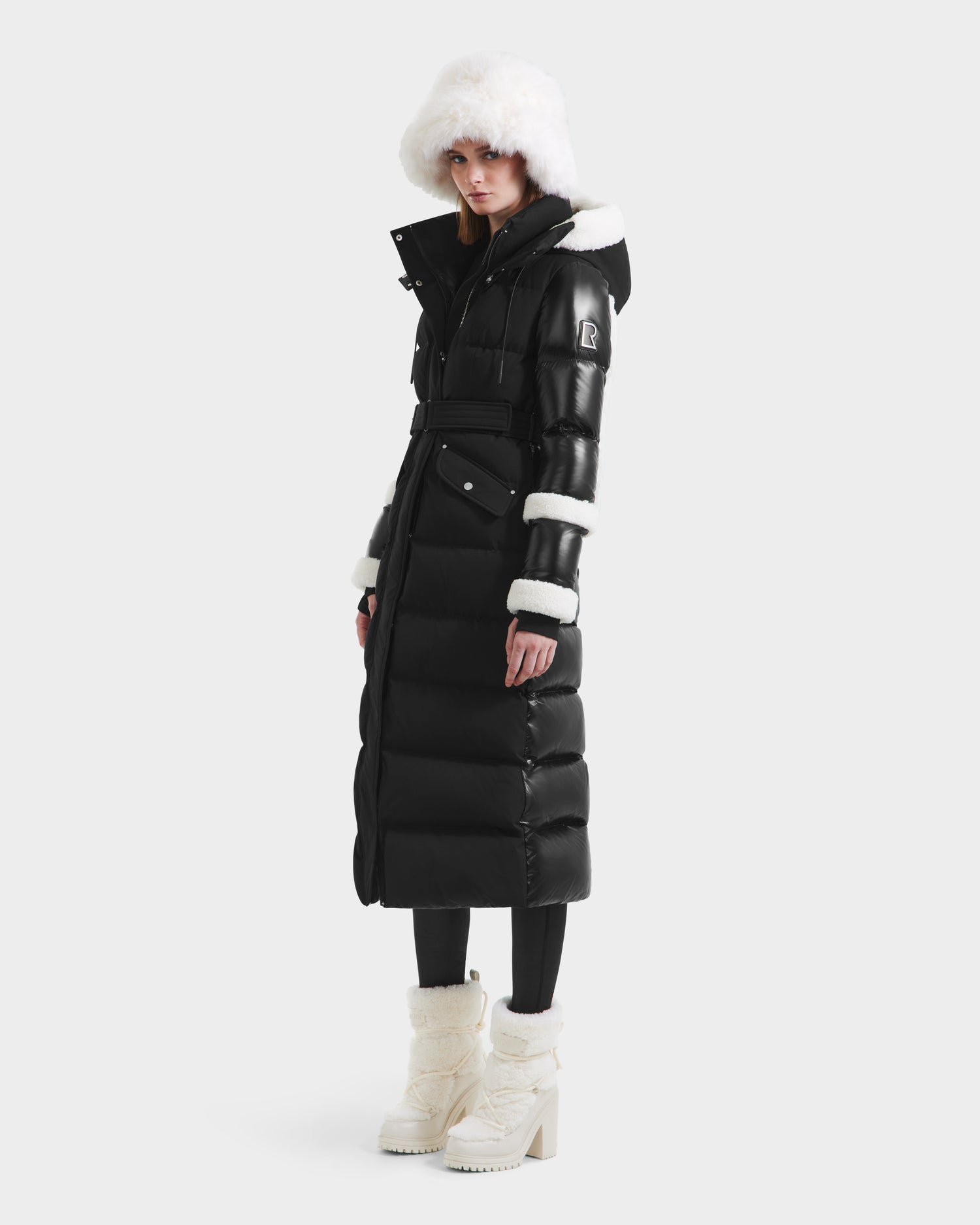 A woman wears the Rudsak AUDREY XL - 8125601 Black/Iceland maxi-length down puffer, featuring faux shearling on the hood, cuffs, and collar, styled with black leggings and white heeled winter boots against a plain light backdrop.