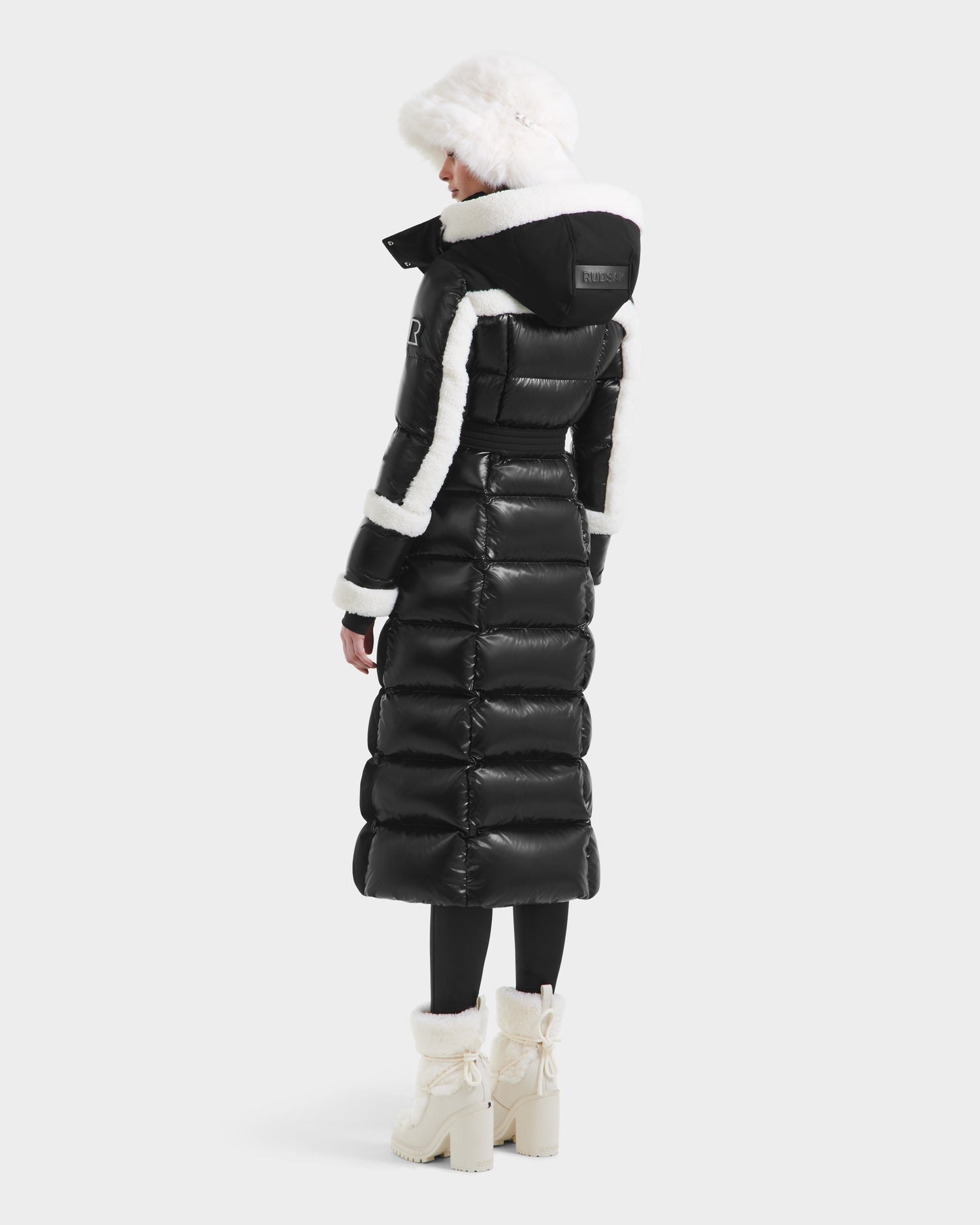 A person stands facing away, wearing the Rudsak AUDREY XL - 8125601 Black/Iceland maxi down puffer with faux shearling trim, a white fur hat, black leggings, and white fur-lined boots against a plain light background.