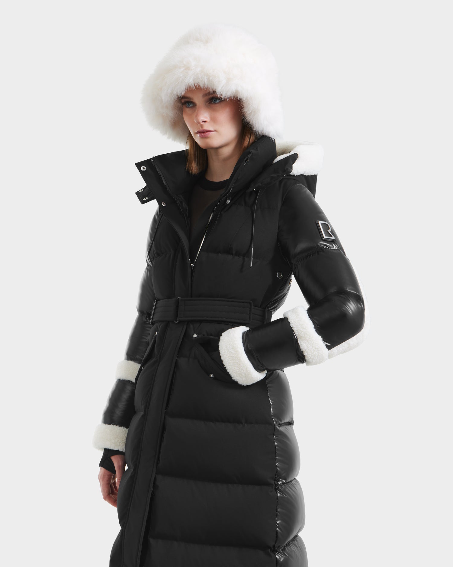 A woman models the Rudsak AUDREY XL - 8125601 Black/Iceland maxi-length down puffer, featuring a belted waist, premium insulation, and white fur trim on the hood, cuffs, and collar with a classic quilted design.