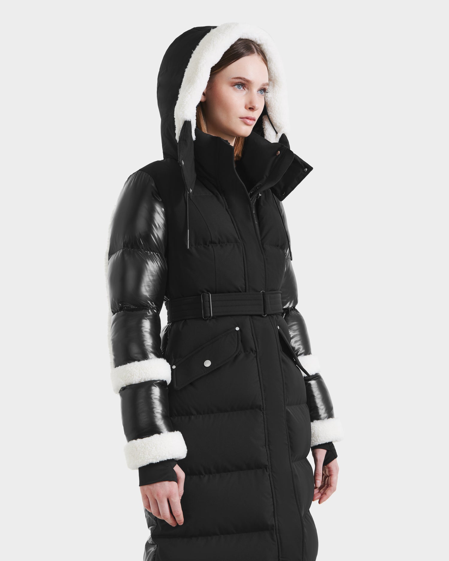 A woman models the Rudsak AUDREY XL - 8125601 Black/Iceland maxi down puffer with a belted waist, glossy sleeves, and white faux shearling trim on the hood and cuffs, standing against a plain light background.