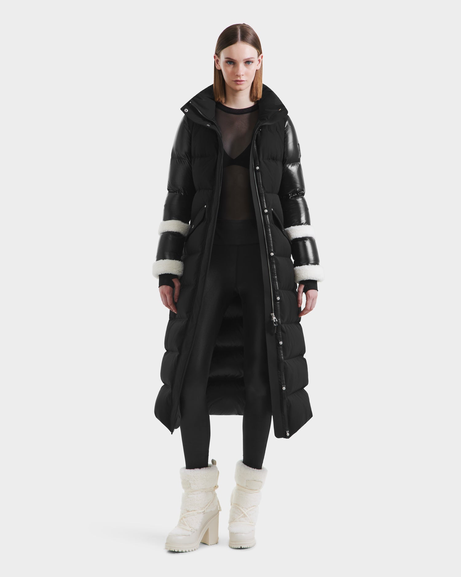 A woman stands facing forward in the Rudsak AUDREY XL - 8125601 Black/Iceland maxi down puffer with white sleeve accents, styled with black leggings, a sheer black top, and white heeled boots against a plain white background.