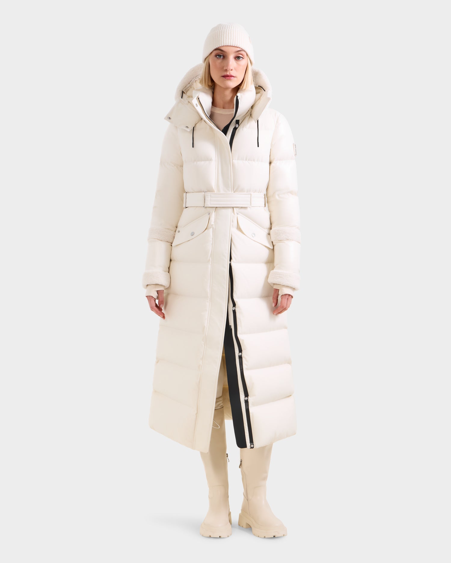 A person stands against a plain light gray background wearing the Rudsak AUDREY XL - 8125601 Vanilla cream maxi-length down puffer jacket with a belt, paired with a matching beanie and cream boots.