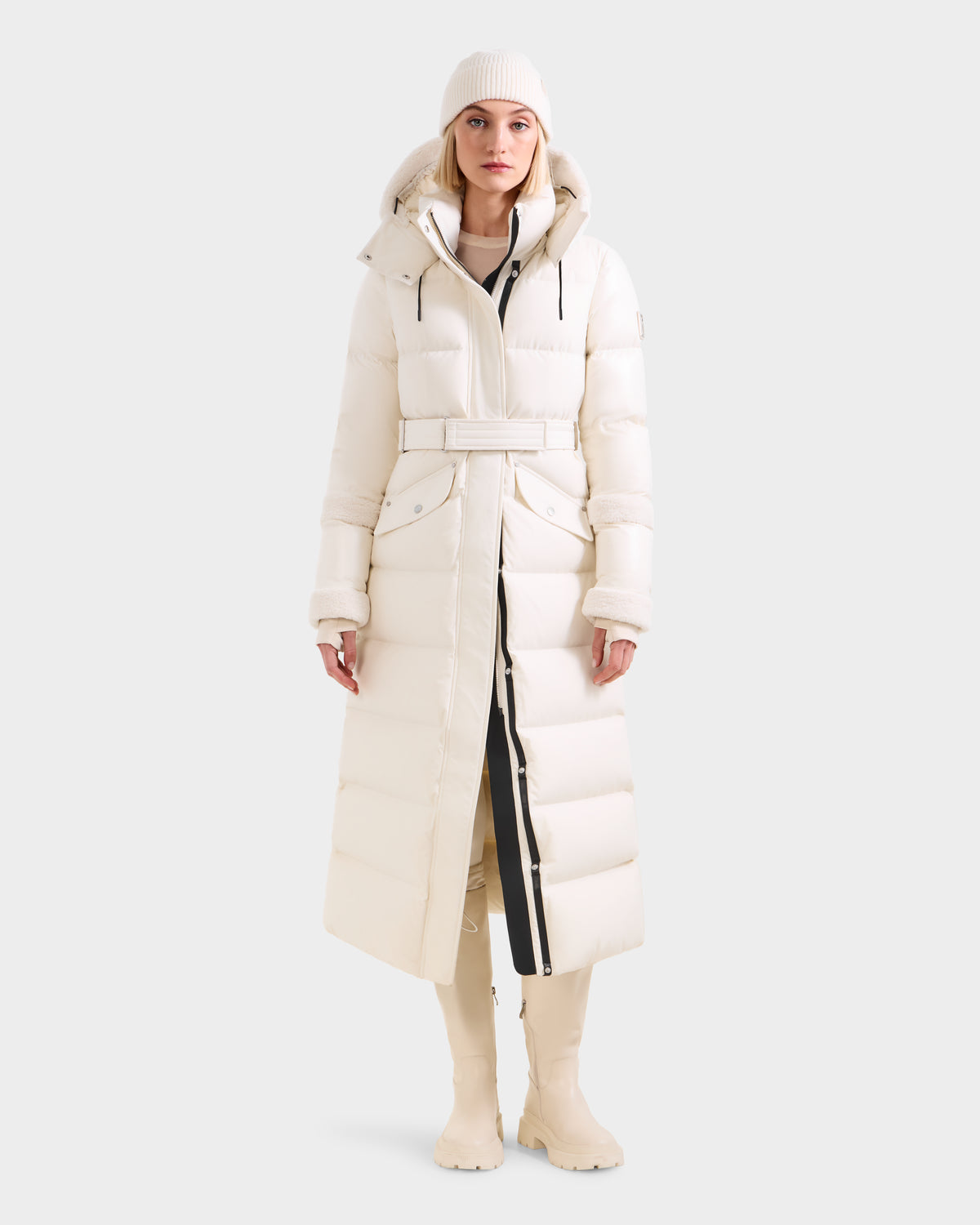 A person stands against a plain light gray background wearing the Rudsak AUDREY XL - 8125601 Vanilla cream maxi-length down puffer jacket with a belt, paired with a matching beanie and cream boots.