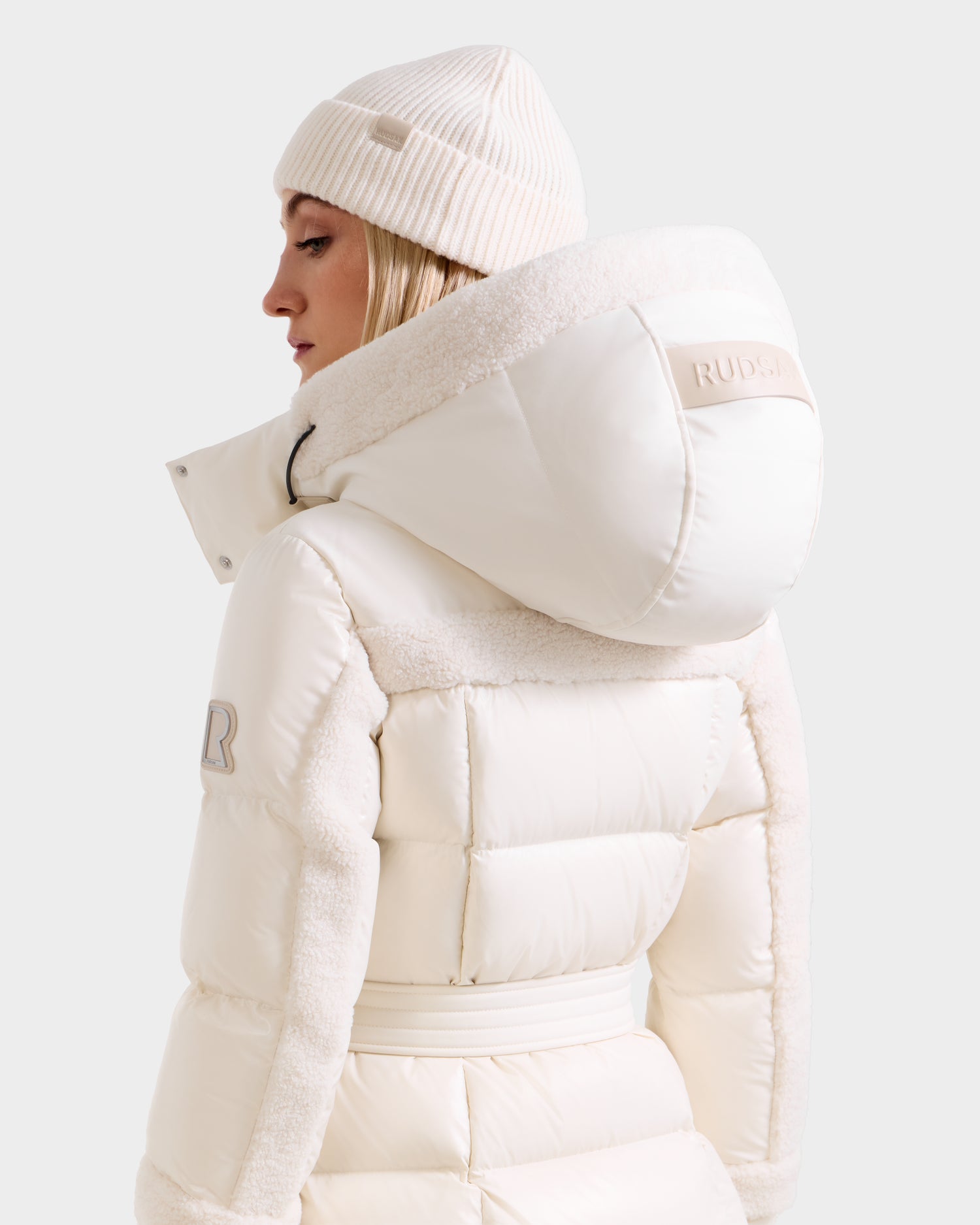 A woman wears the Rudsak AUDREY XL - 8125601 Vanilla, a cream maxi down puffer with a large hood and matching knit beanie. "RUDSAK" is displayed on the back with textured panels. The background is plain white.
