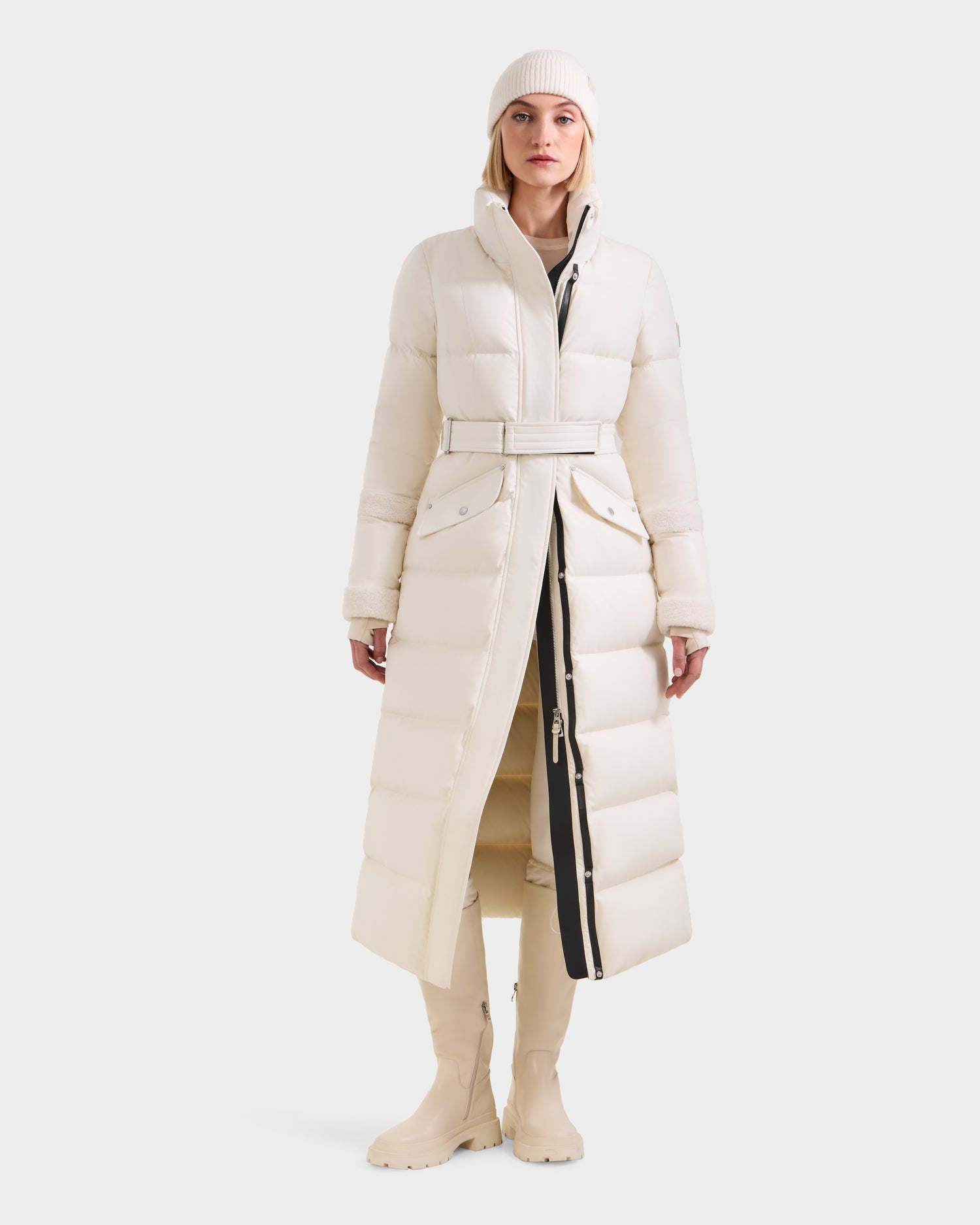 A woman faces forward, modeling the Rudsak AUDREY XL - 8125601 Vanilla maxi down puffer jacket with a belt, paired with tall matching boots, neutral pants, and a white knit beanie against a plain light background.