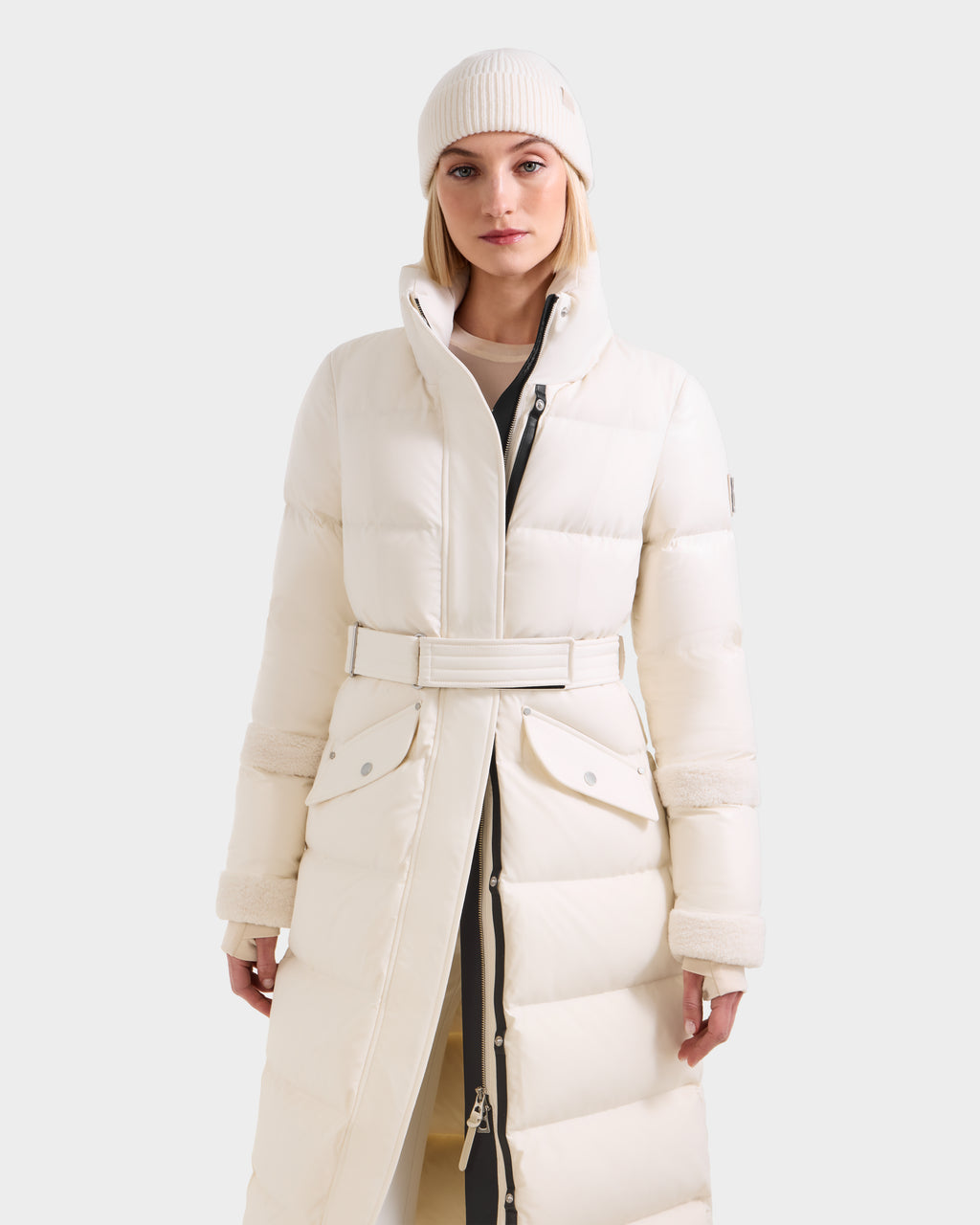 A woman faces forward against a plain background, wearing the Rudsak AUDREY XL - 8125601 Vanilla maxi-length down puffer jacket with a belted waist and a matching white knit beanie for a sleek silhouette.