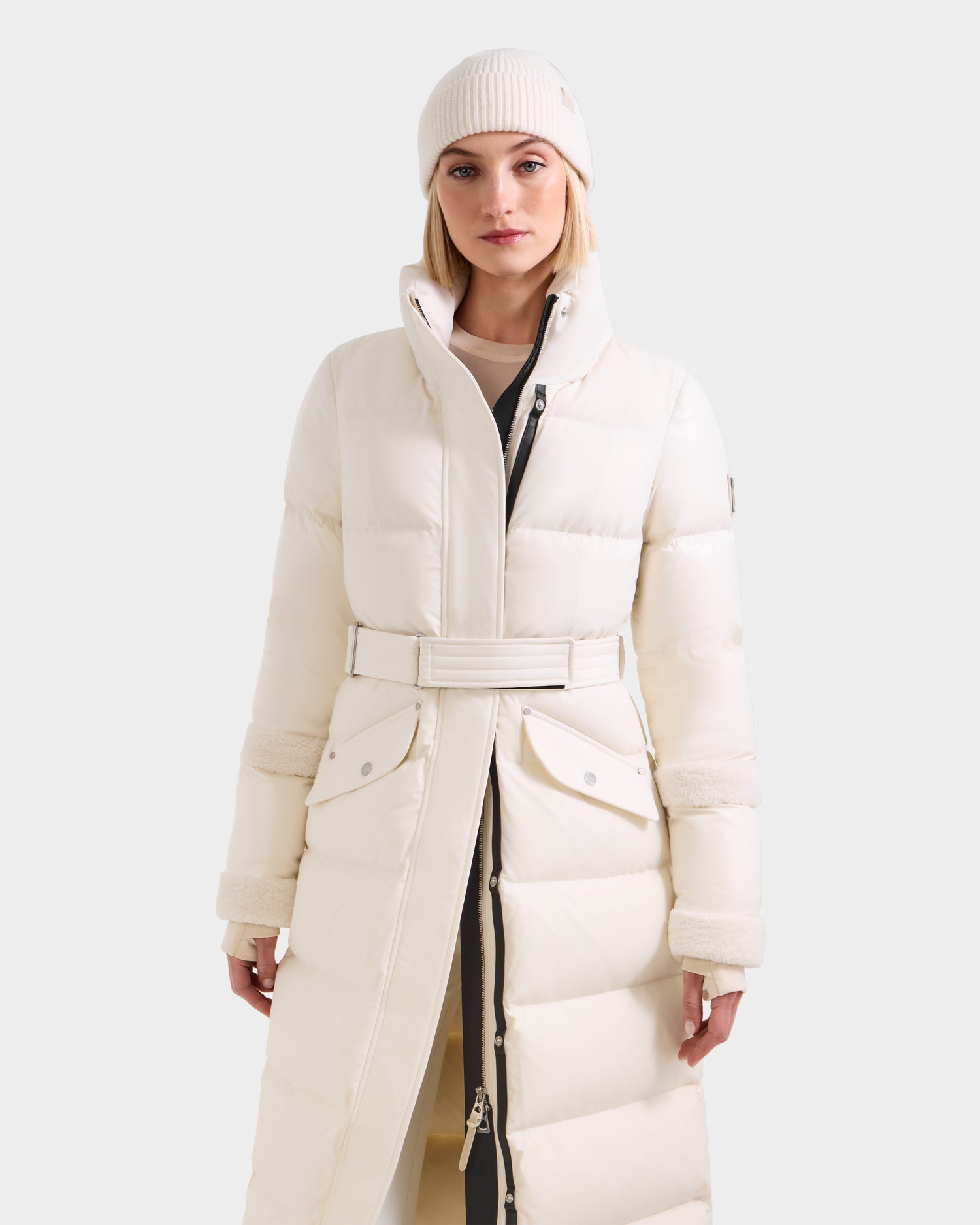 A woman faces forward against a plain background, wearing the Rudsak AUDREY XL - 8125601 Vanilla maxi-length down puffer jacket with a belted waist and a matching white knit beanie for a sleek silhouette.