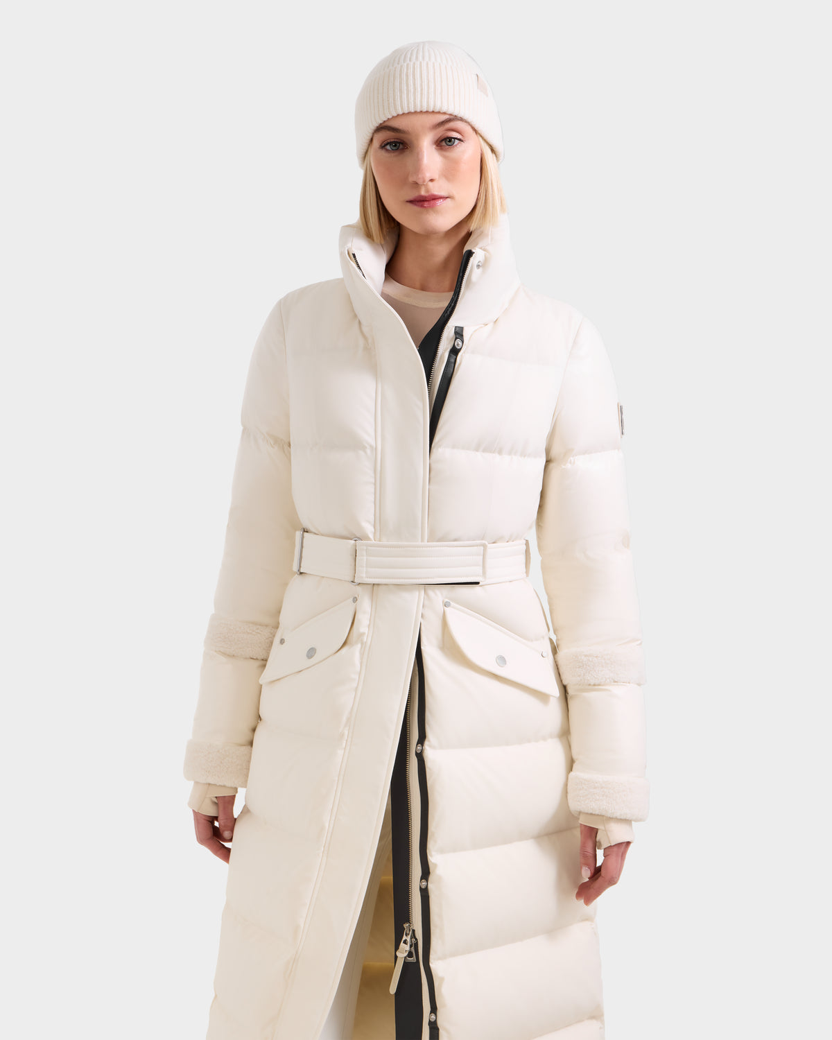 A woman faces forward against a plain background, wearing the Rudsak AUDREY XL - 8125601 Vanilla maxi-length down puffer jacket with a belted waist and a matching white knit beanie for a sleek silhouette.