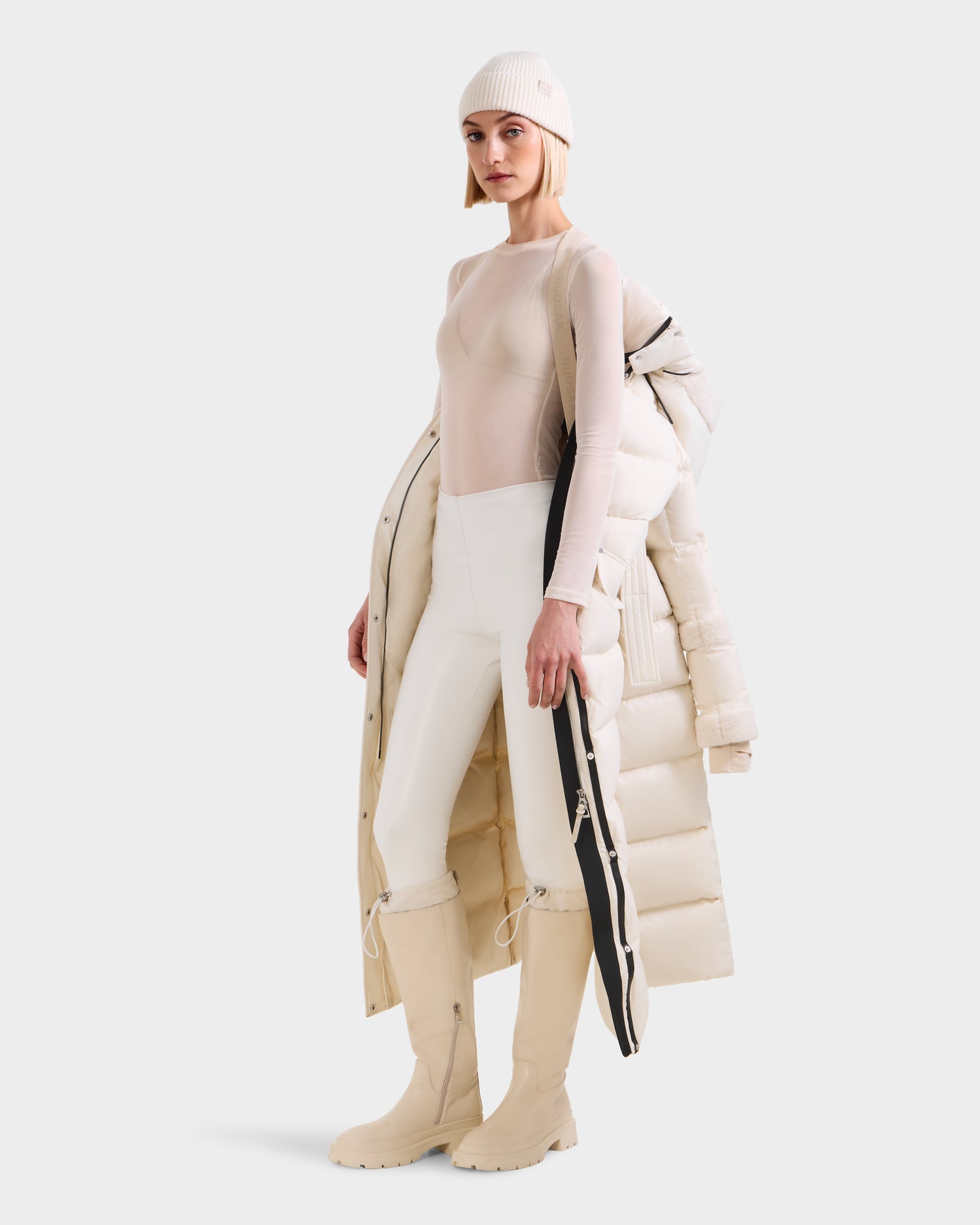 A woman in a beige beanie, sheer top, white pants, and knee-high boots poses with the Rudsak AUDREY XL - 8125601 Vanilla maxi-length down puffer jacket draped over one shoulder, showcasing its full silhouette against a plain light background.