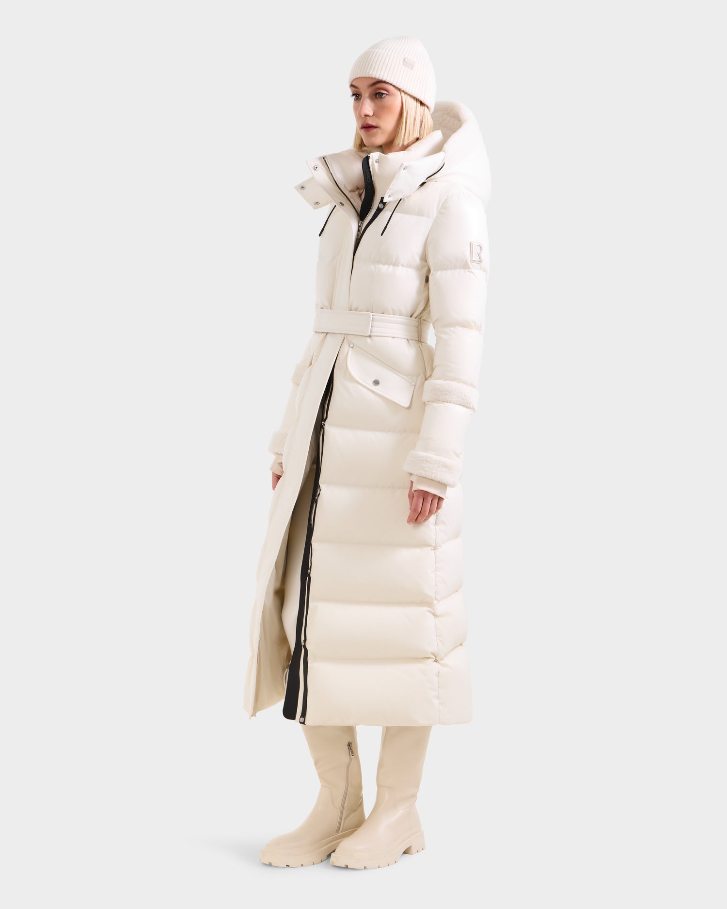 A woman wears the Rudsak AUDREY XL - 8125601 Vanilla maxi-length down puffer jacket with a hood, paired with a white knit beanie and tall white boots, standing against a plain light background.