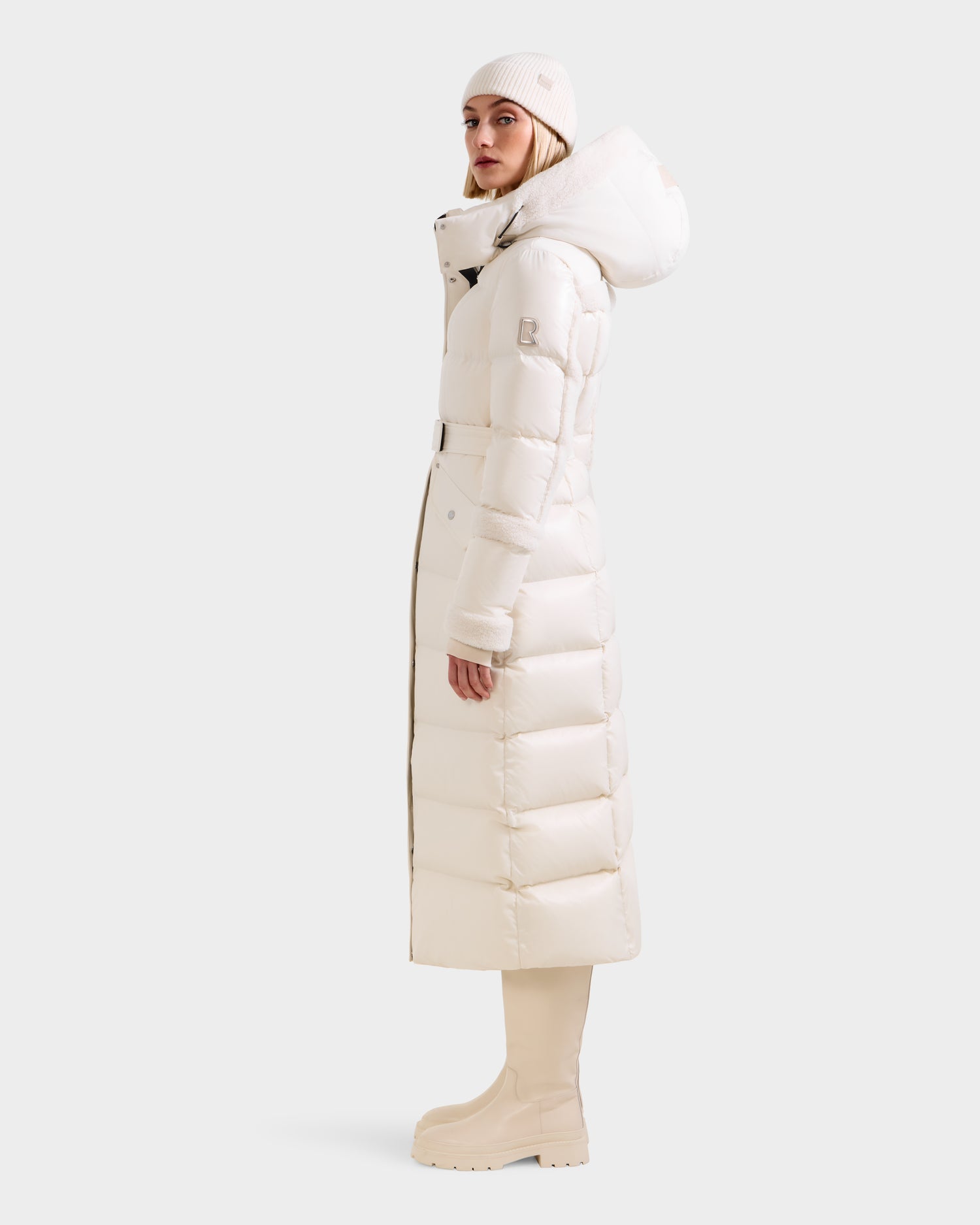 A person models the Rudsak AUDREY XL - 8125601 Vanilla maxi-length down puffer jacket with hood, paired with a white hat and cream boots, standing sideways against a plain light background.