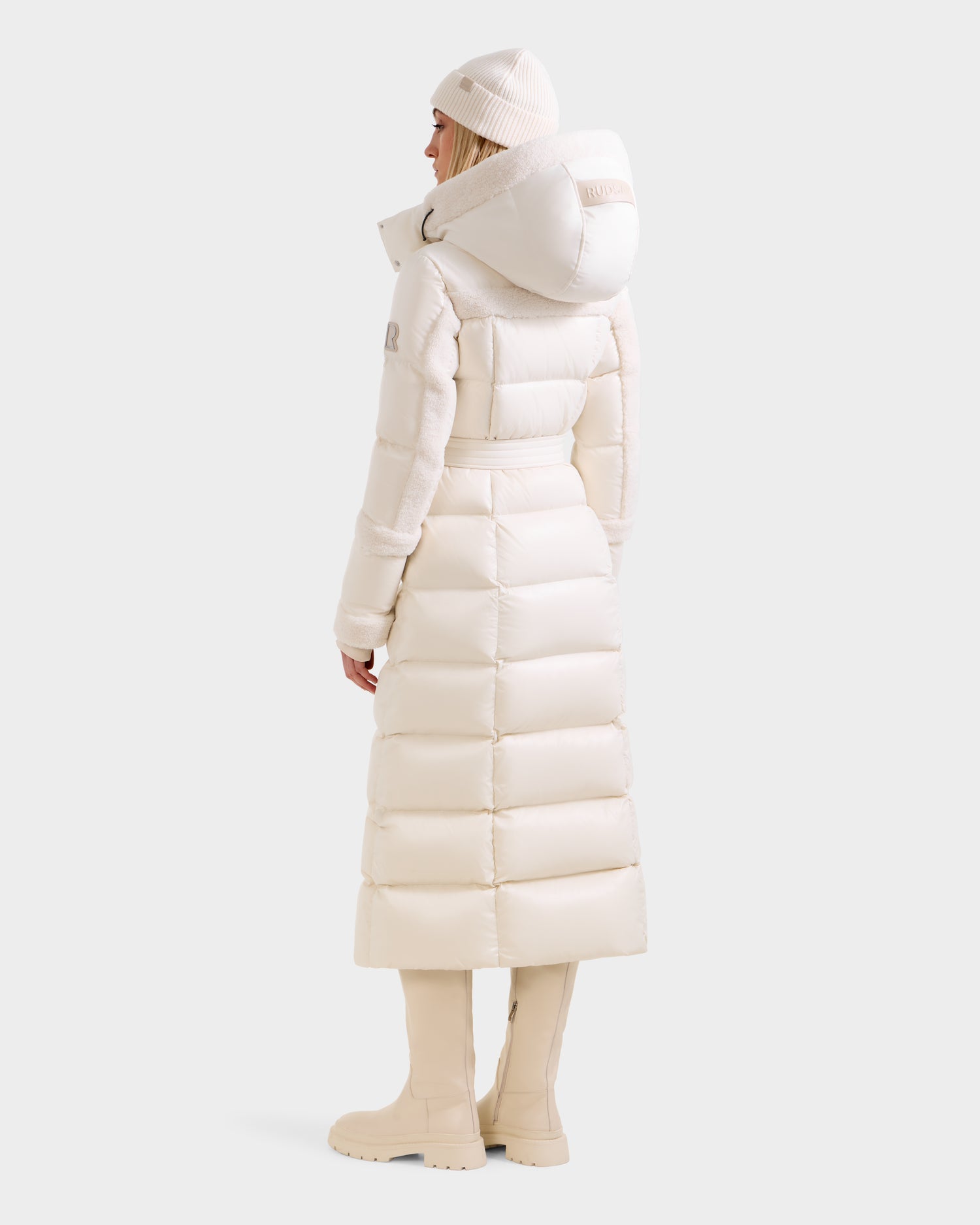A person faces away in a Rudsak AUDREY XL - 8125601 Vanilla maxi down puffer with a large hood, cream beanie, and knee-high boots against a plain light background.