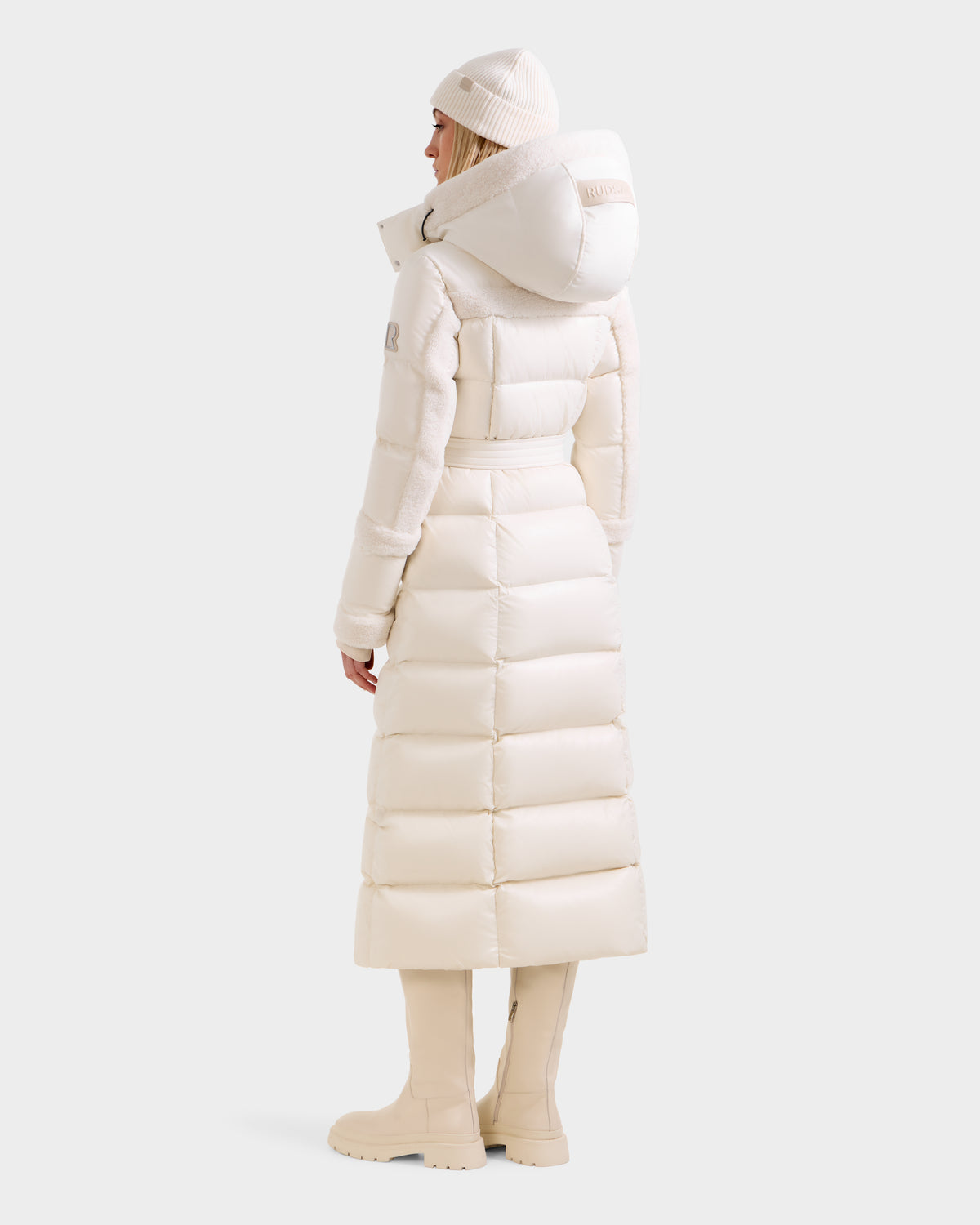 A person faces away in a Rudsak AUDREY XL - 8125601 Vanilla maxi down puffer with a large hood, cream beanie, and knee-high boots against a plain light background.