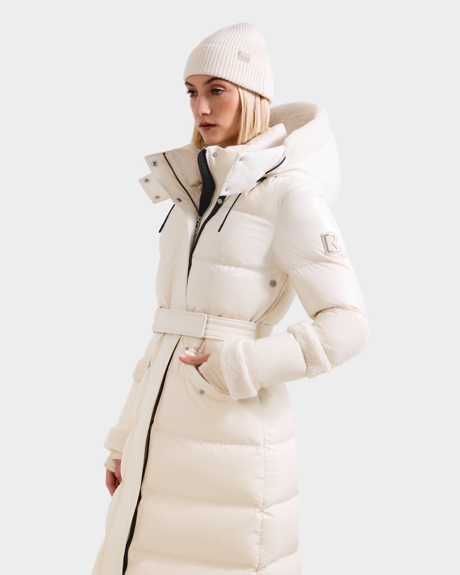 A woman wears the Rudsak AUDREY XL - 8125601 Vanilla maxi down puffer with high collar and hood, made from recycled Stormshell fabric, paired with a white knit beanie. She stands against a light background, looking to the side with one hand in her pocket.