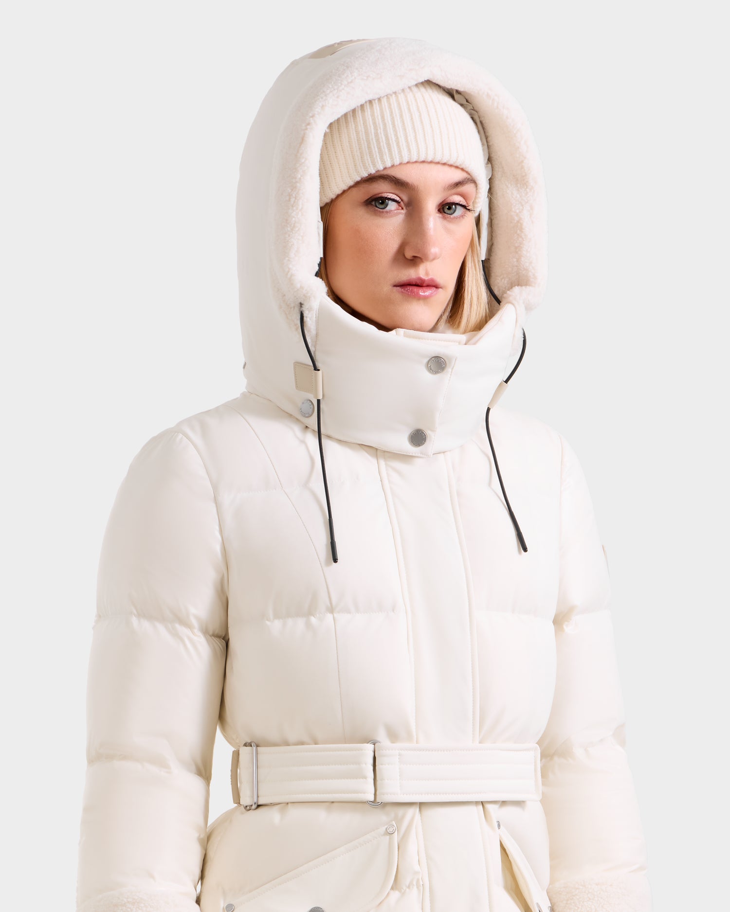 A woman models the Rudsak AUDREY XL - 8125601 Vanilla, a white maxi-length down puffer with belted waist and faux fur-lined hood, paired with a cream knit beanie against a plain light background.