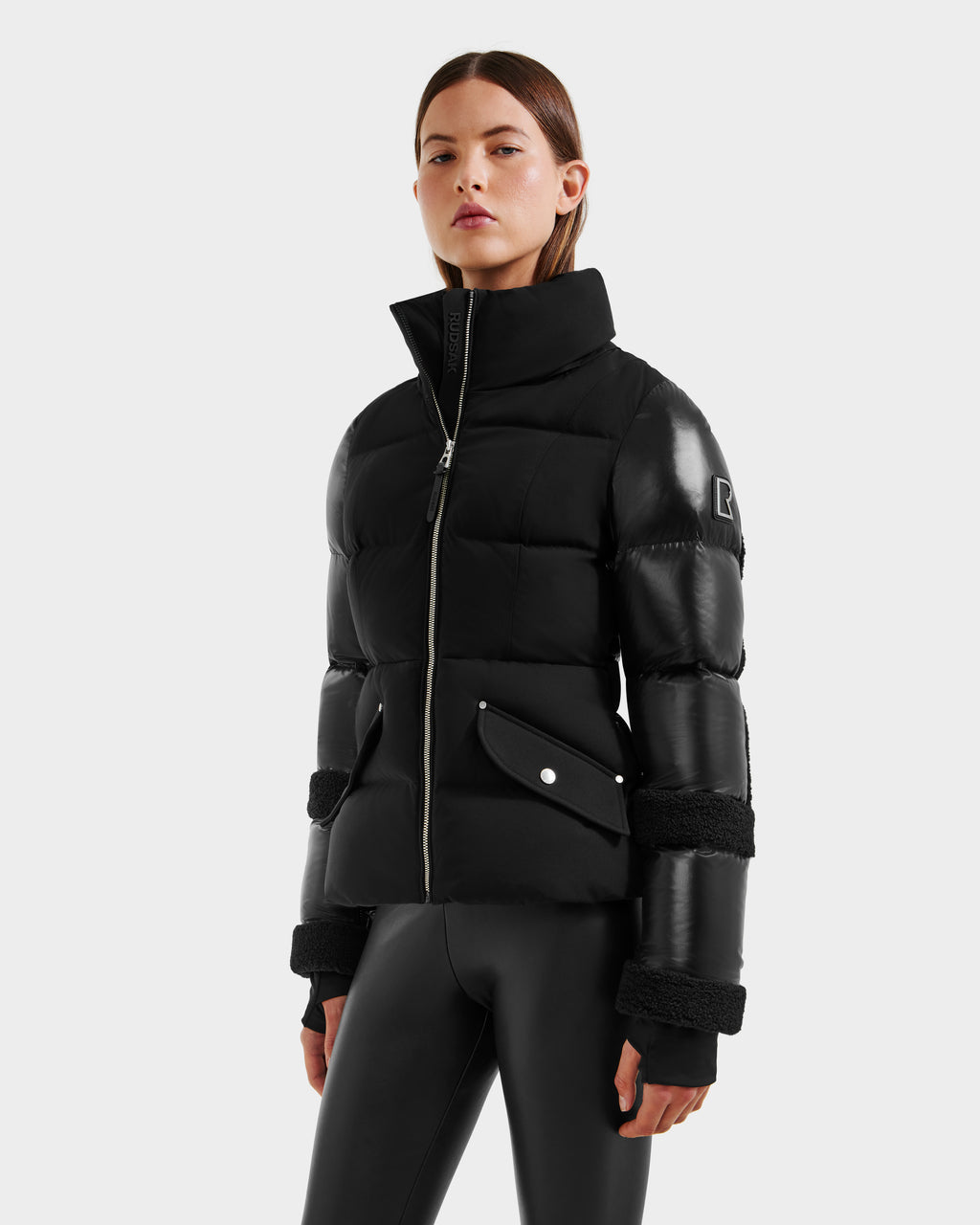 A woman wears the Rudsak AUDREY - 8124582 Black/Black down puffer jacket with leather sleeves, crafted from recycled Stormshell fabric, and shiny black leggings, against a light gray background. The jacket has a high collar and silver zipper.