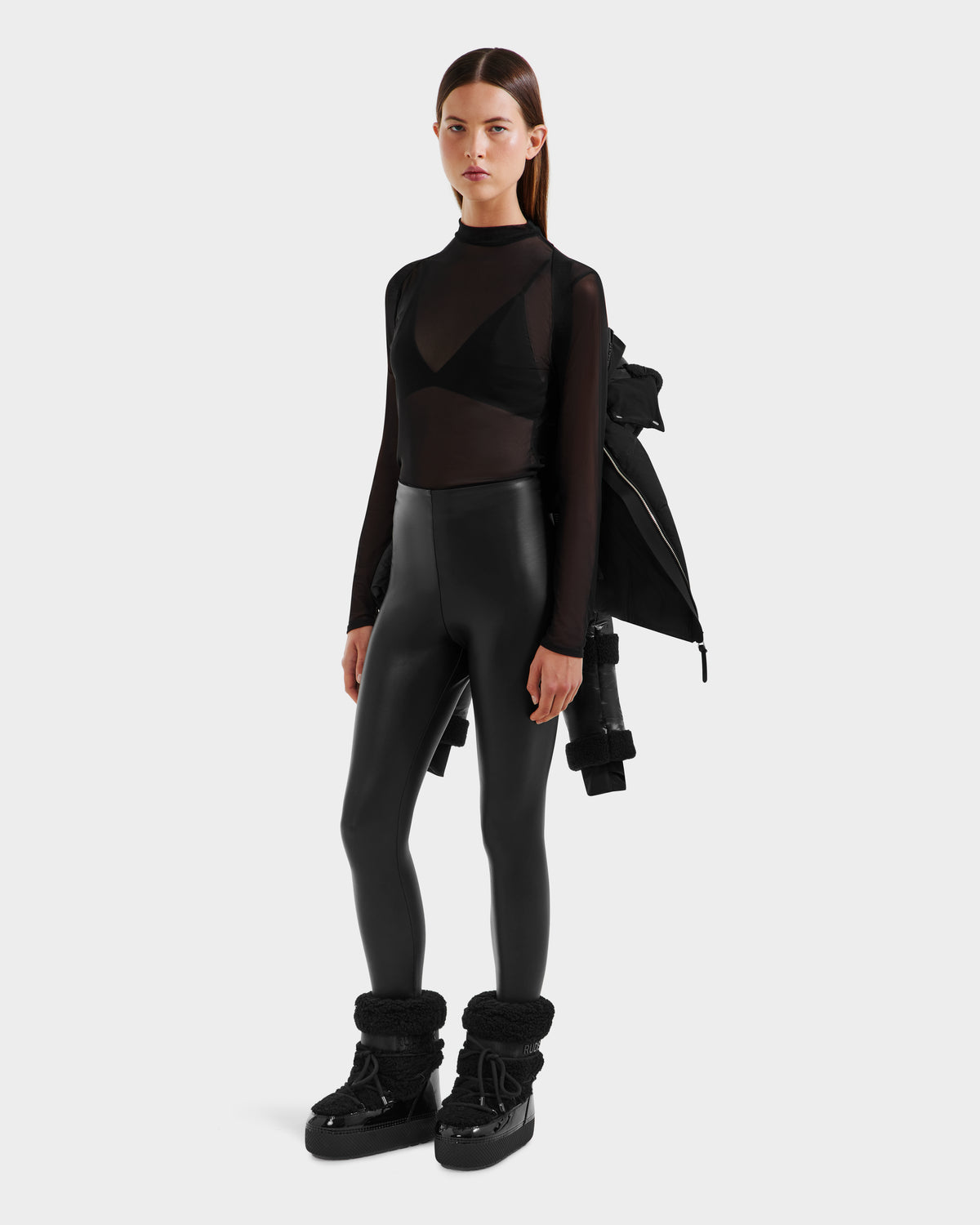 A woman stands against a plain background wearing the Rudsak AUDREY - 8124582 Black/Black down puffer jacket with RDS certified down, paired with a sheer black top, black bra, leggings, and chunky boots.