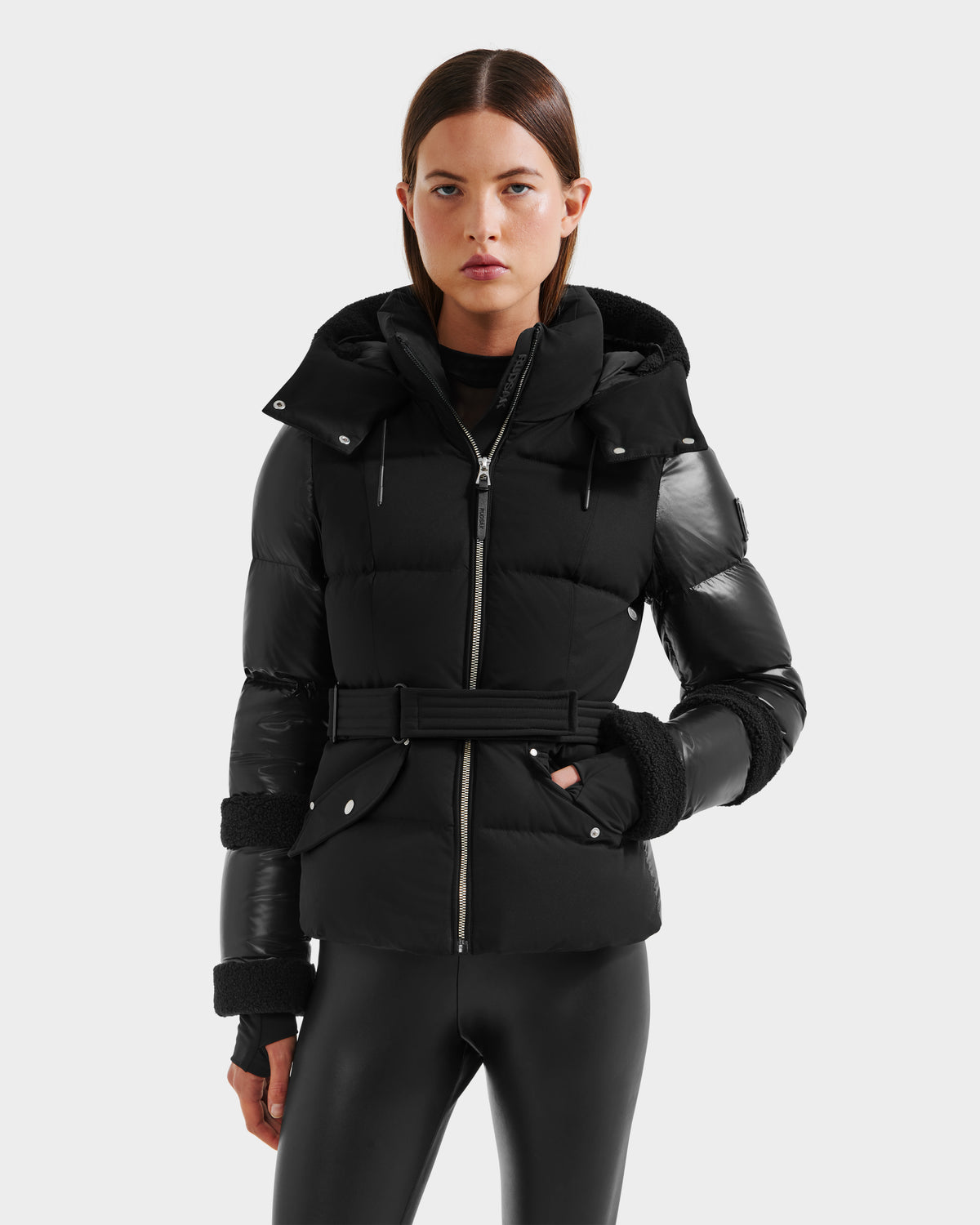 A woman wears the Rudsak AUDREY - 8124582 Black/Black down puffer jacket with hood and belted waist, paired with black leggings, standing facing forward against a plain light gray background.