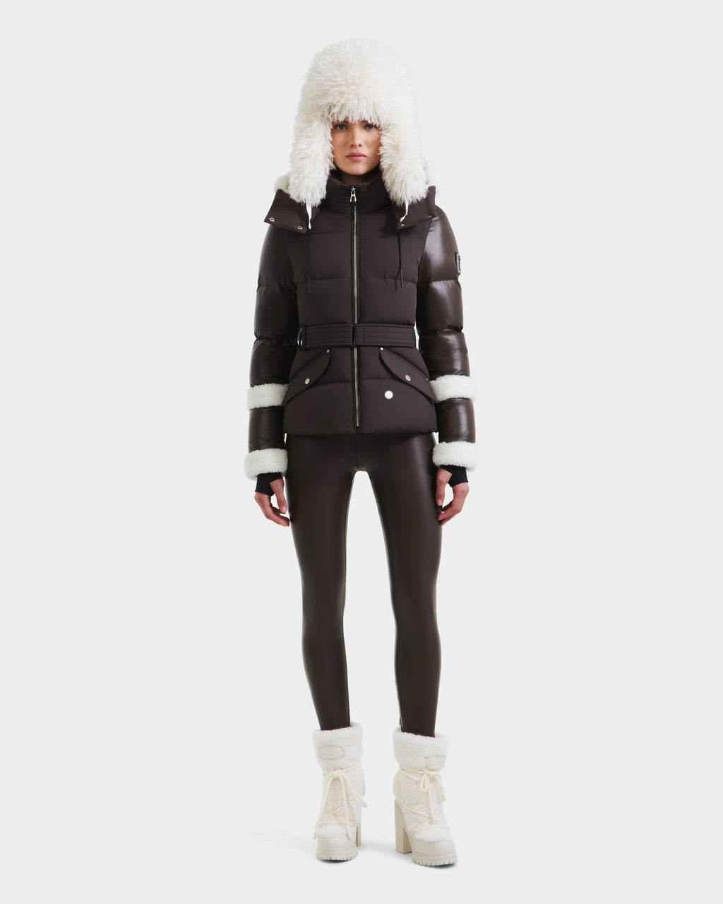A person faces forward against a white background, wearing the Rudsak AUDREY - 8124582 Espresso puffer jacket with recycled Stormshell fabric and white fur trim, paired with matching dark leggings and white fur-lined boots.