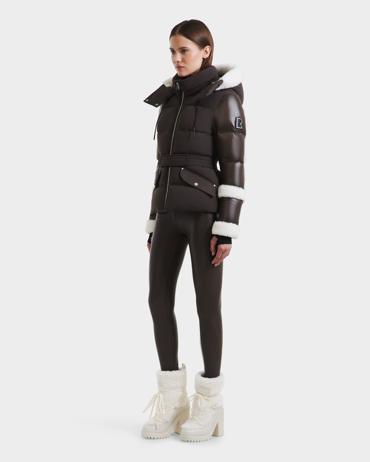 A woman models the Rudsak AUDREY - 8124582 Espresso brown down puffer jacket with white faux fur trim, paired with matching leggings and fur-lined boots against a plain white background.