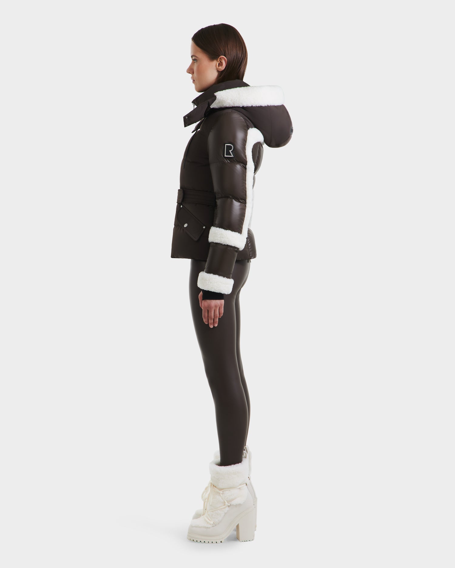 A woman stands in profile wearing the Rudsak AUDREY - 8124582 Espresso down puffer jacket, made from recycled Stormshell fabric with white fur trim and hood. She pairs it with black leggings and white platform boots; a patch details the sleeve.