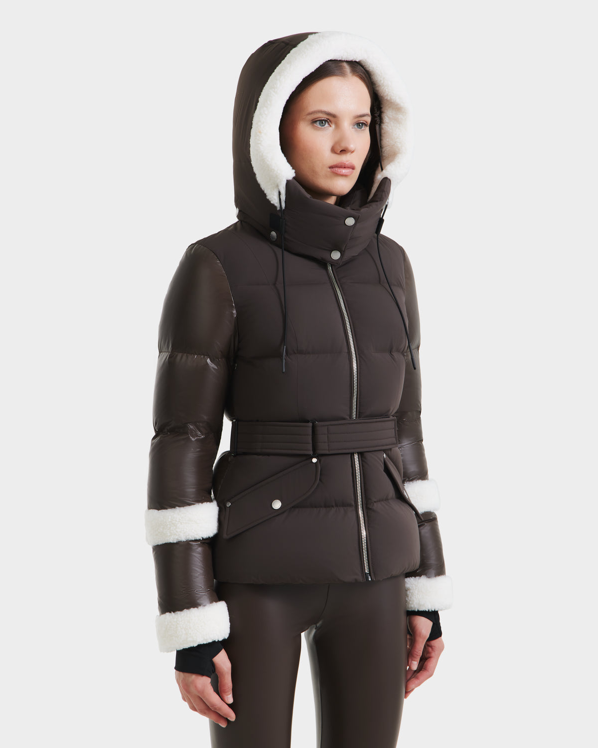 A woman wears the Rudsak AUDREY - 8124582 Espresso dark brown down puffer jacket with white faux fur trim and a wide waist belt. Filled with RDS certified down, it stands out against a plain white background.