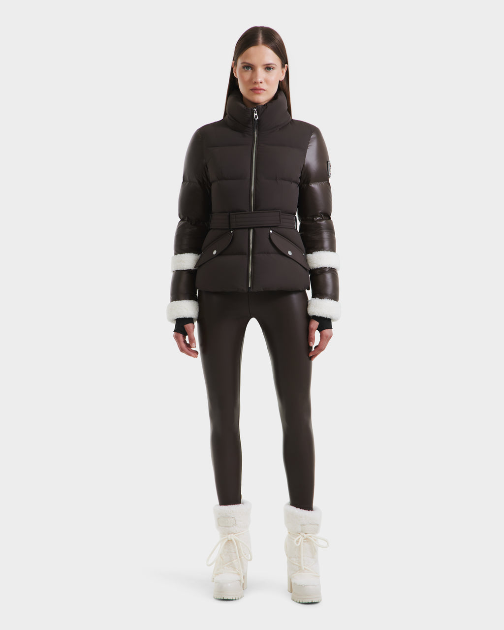 A woman stands facing forward in a Rudsak AUDREY - 8124582 Espresso down puffer jacket with white fur trim on the sleeves, black leggings, and white fur-lined winter boots against a plain white background.