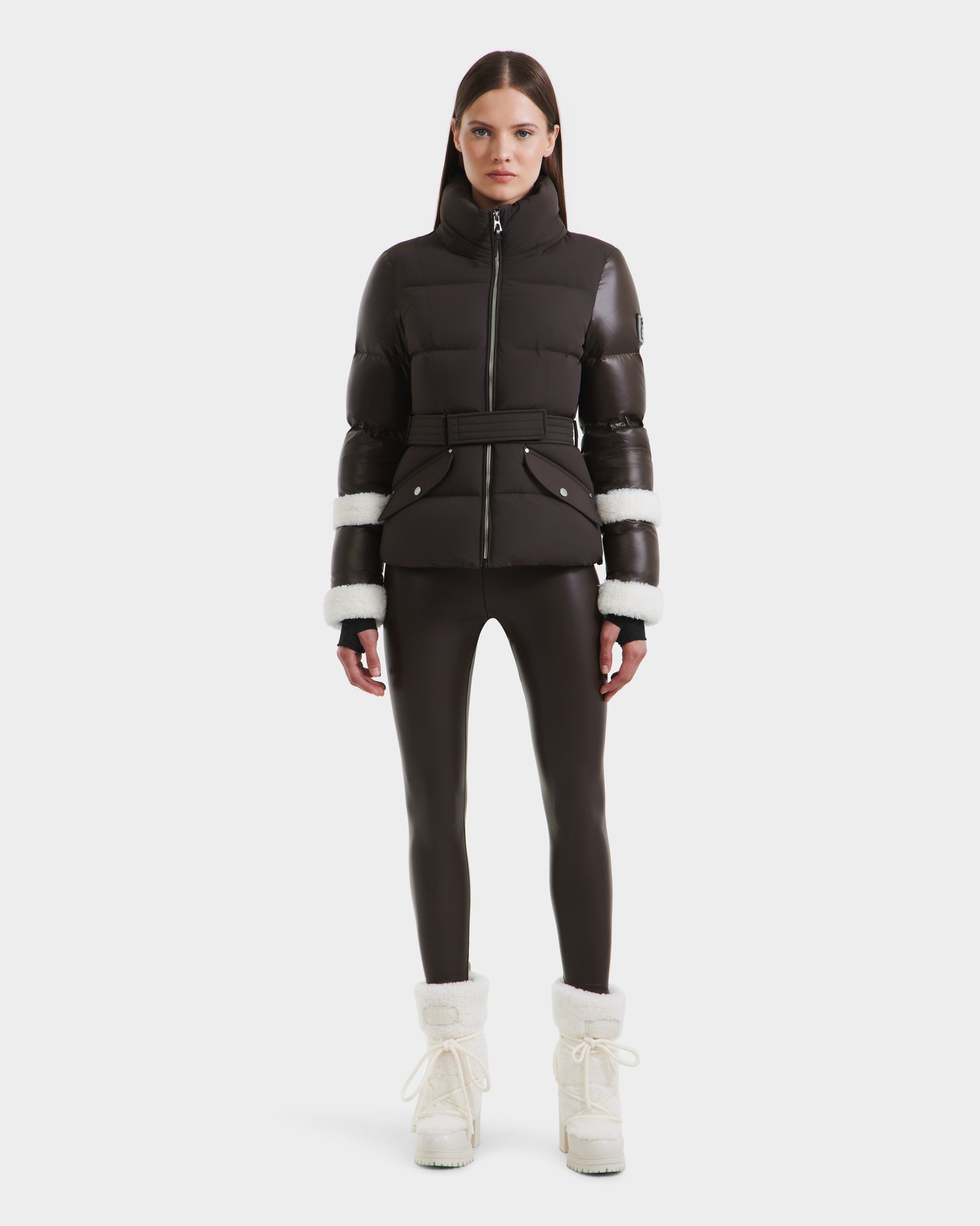 A woman stands facing forward in a Rudsak AUDREY - 8124582 Espresso down puffer jacket with white fur trim on the sleeves, black leggings, and white fur-lined winter boots against a plain white background.