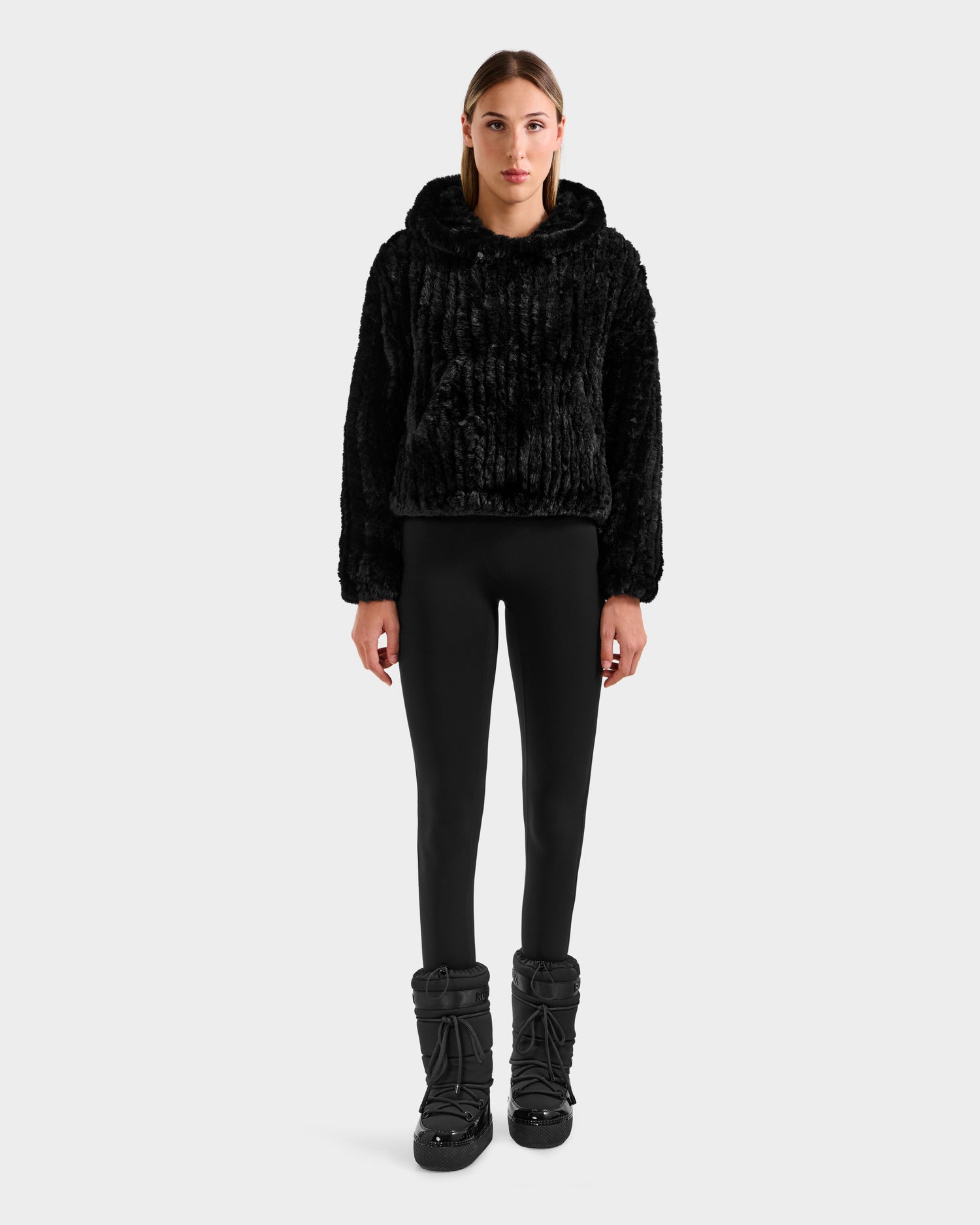 A person faces forward, dressed for winter lounging in the Rudsak AUGUST - 8525658 Black faux fur hoodie, black leggings, and black winter boots against a plain light gray background.